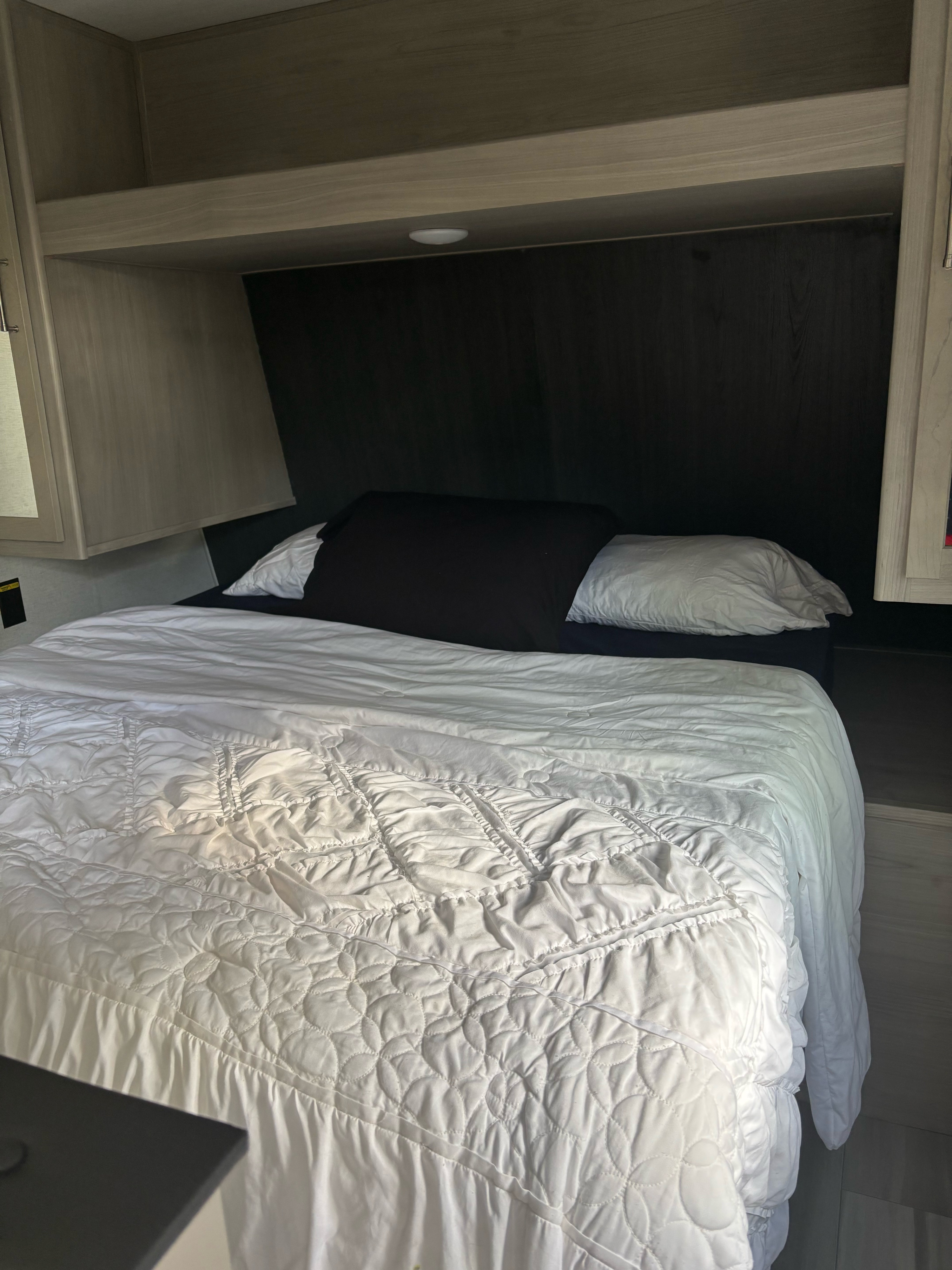 Cozy RV rental bed with white bedding, dark headboard, and wood cabinetry overhead.. Keystone RV Classic 2024