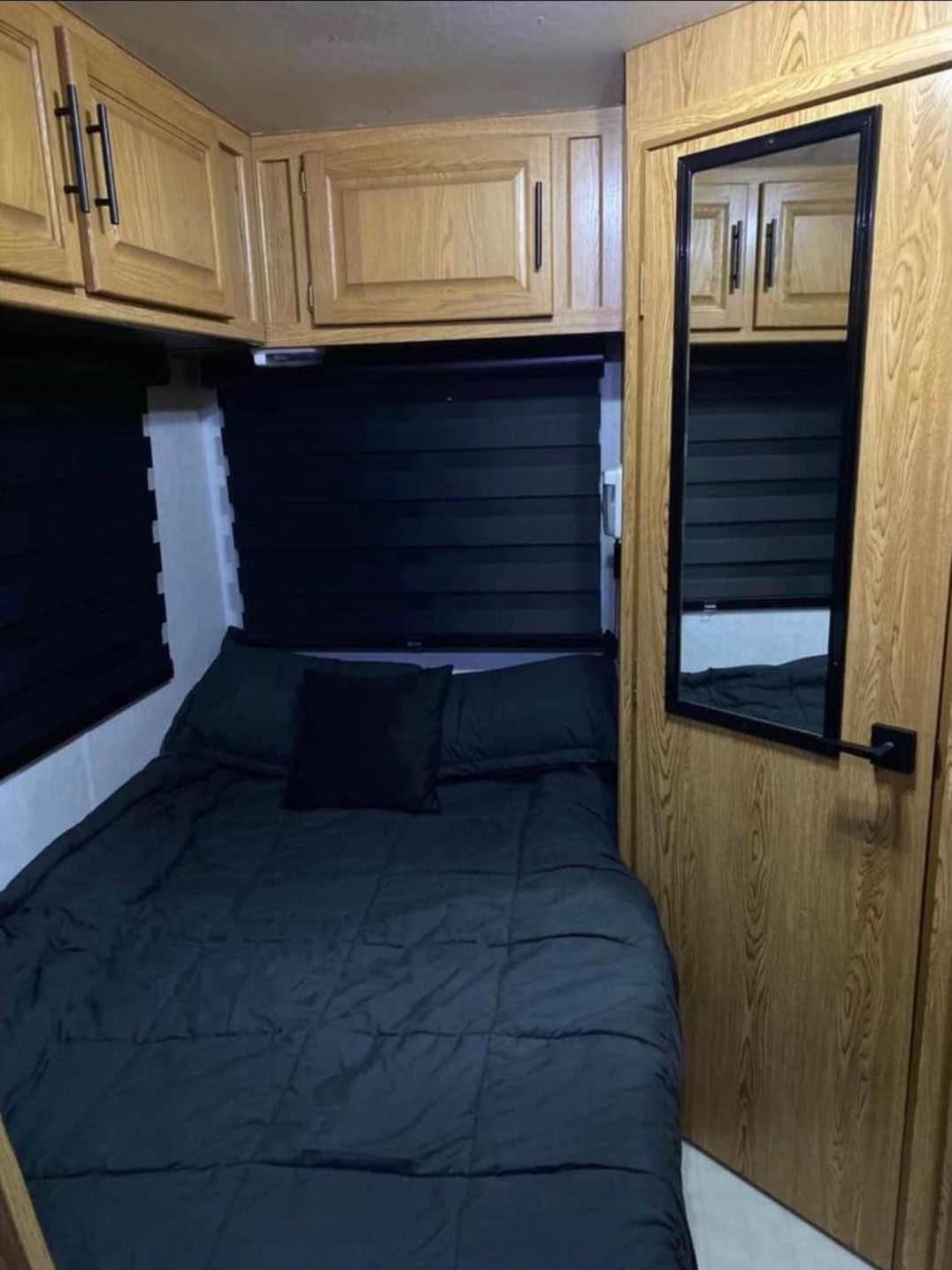 Compact RV rental bedroom featuring a dark bed, overhead wood cabinets, and black window blinds. A mirror on the door aids convenience.. Ford Ford E350 1996