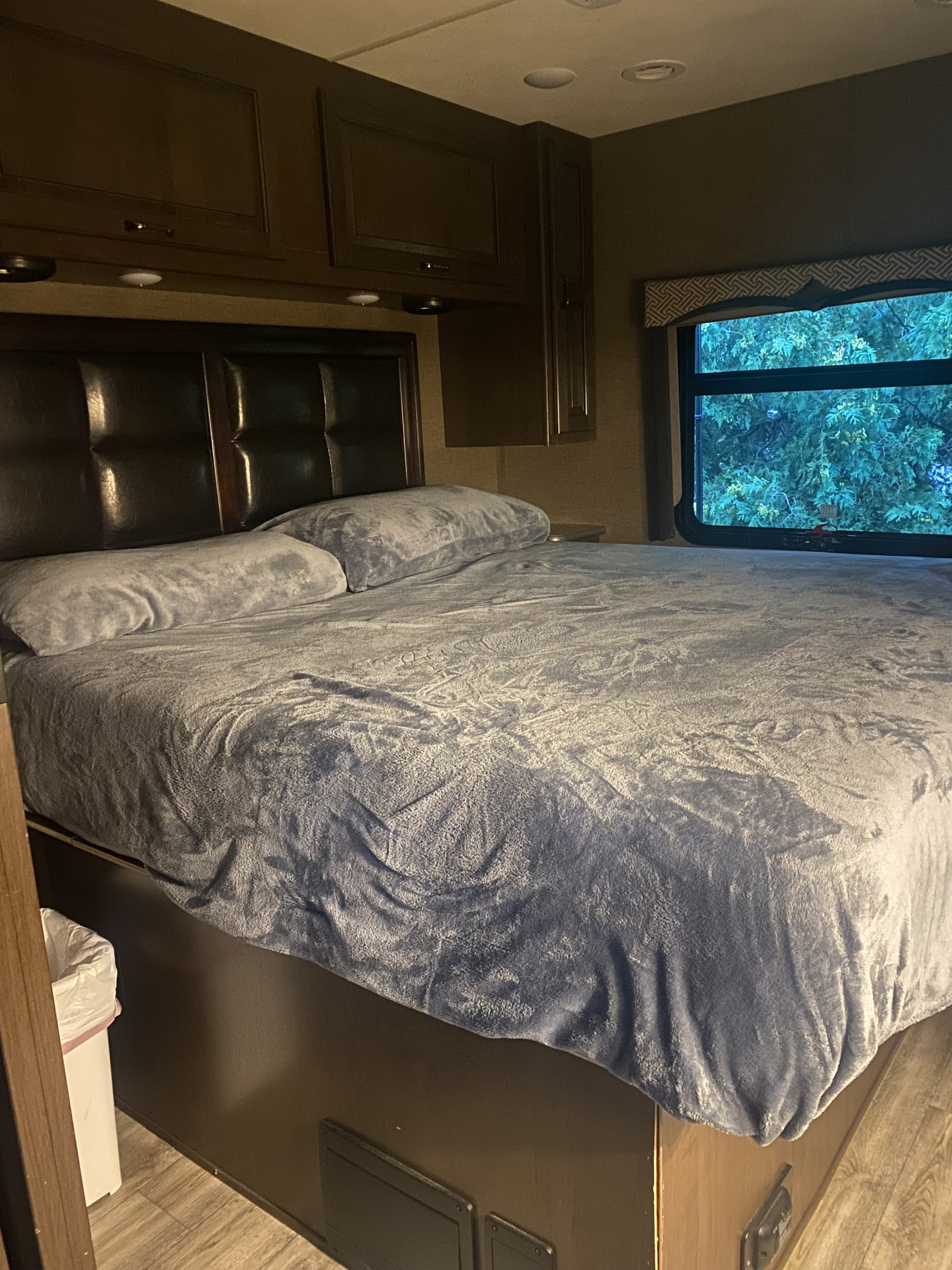 Cozy RV rental bedroom with a large bed, plush gray bedding, dark headboard, overhead cabinets, and a window showing trees outside.. Thor Hurricane 2018
