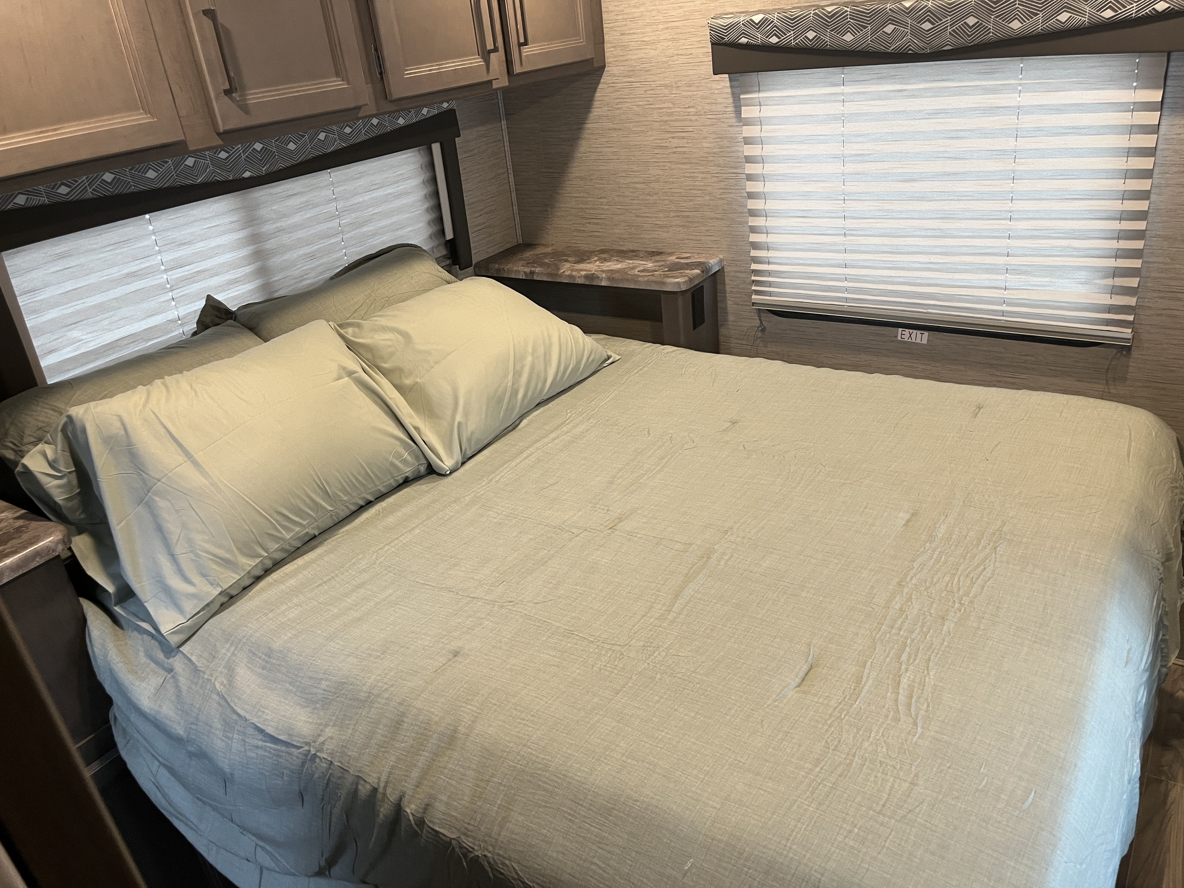 Inside an RV rental bedroom: a made bed with light green bedding, overhead cabinets, and window blinds with an 'EXIT' sign.. Thor Four Winds 2023