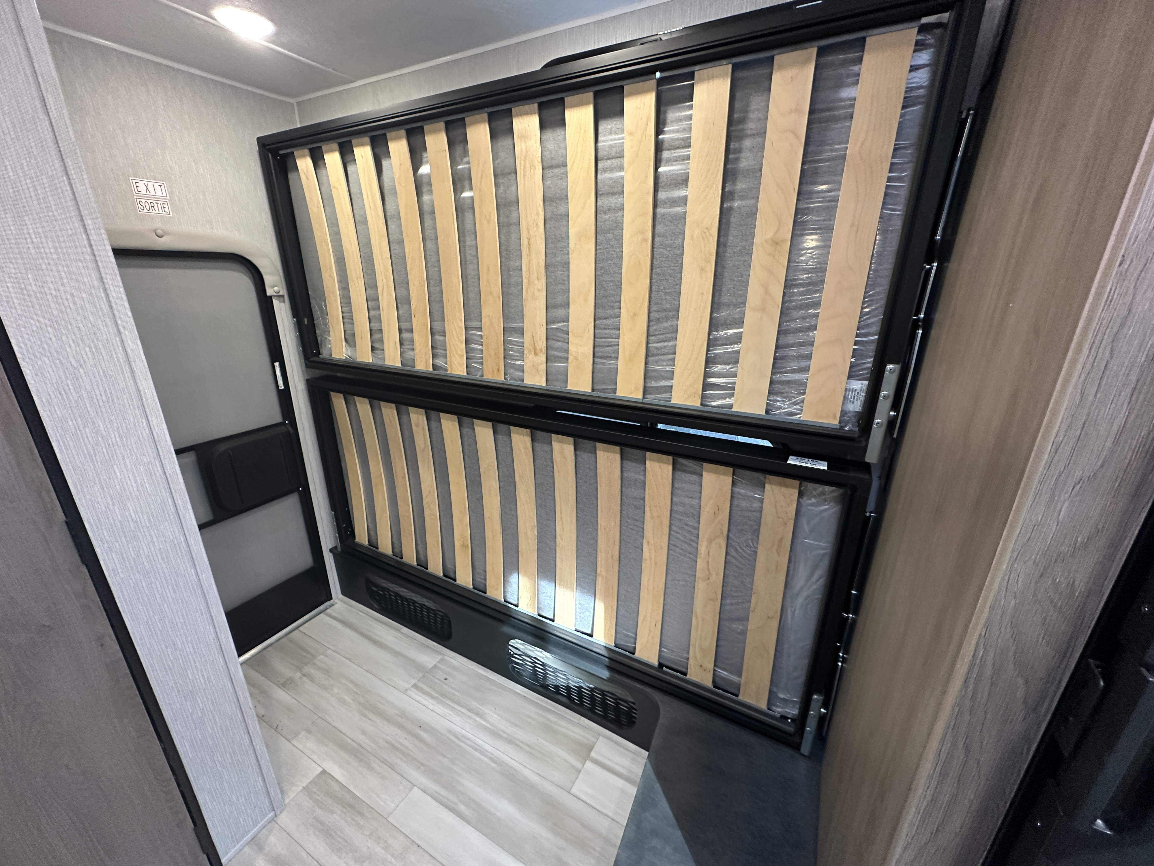 Inside an RV rental: Unused bunk bed frames with wooden slats, still in plastic, beside an exit door. Modern, light interior.. Coachmen Cross Trail 20BHTA 2023
