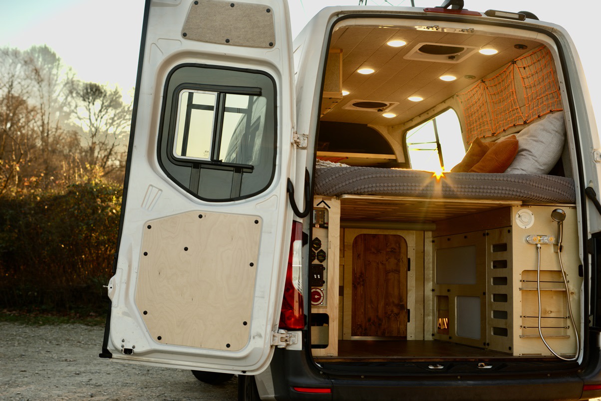 White camper van's rear doors open, showing a custom interior with a cozy bed, lighting, storage, and shower. Perfect for RV rental.. Mercedes-Benz Sprinter 2019