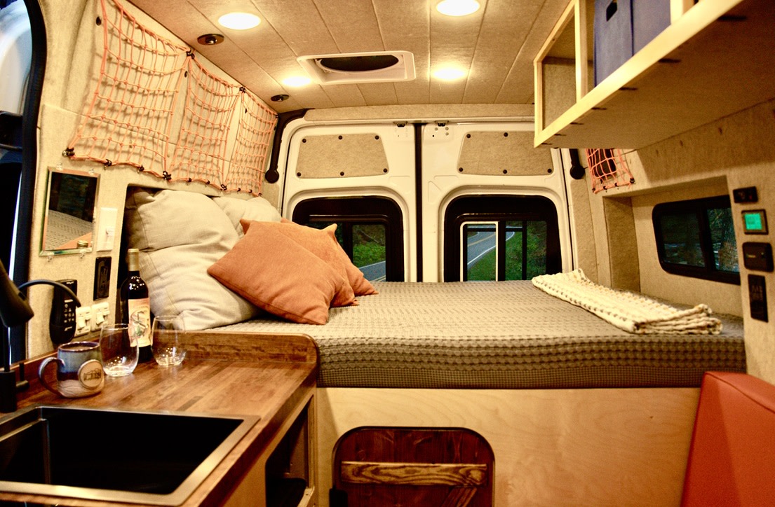Inside this RV rental: a cozy bed, kitchenette with sink, smart storage, and rear windows revealing a road. Van life comfort.. Mercedes-Benz Sprinter 2019