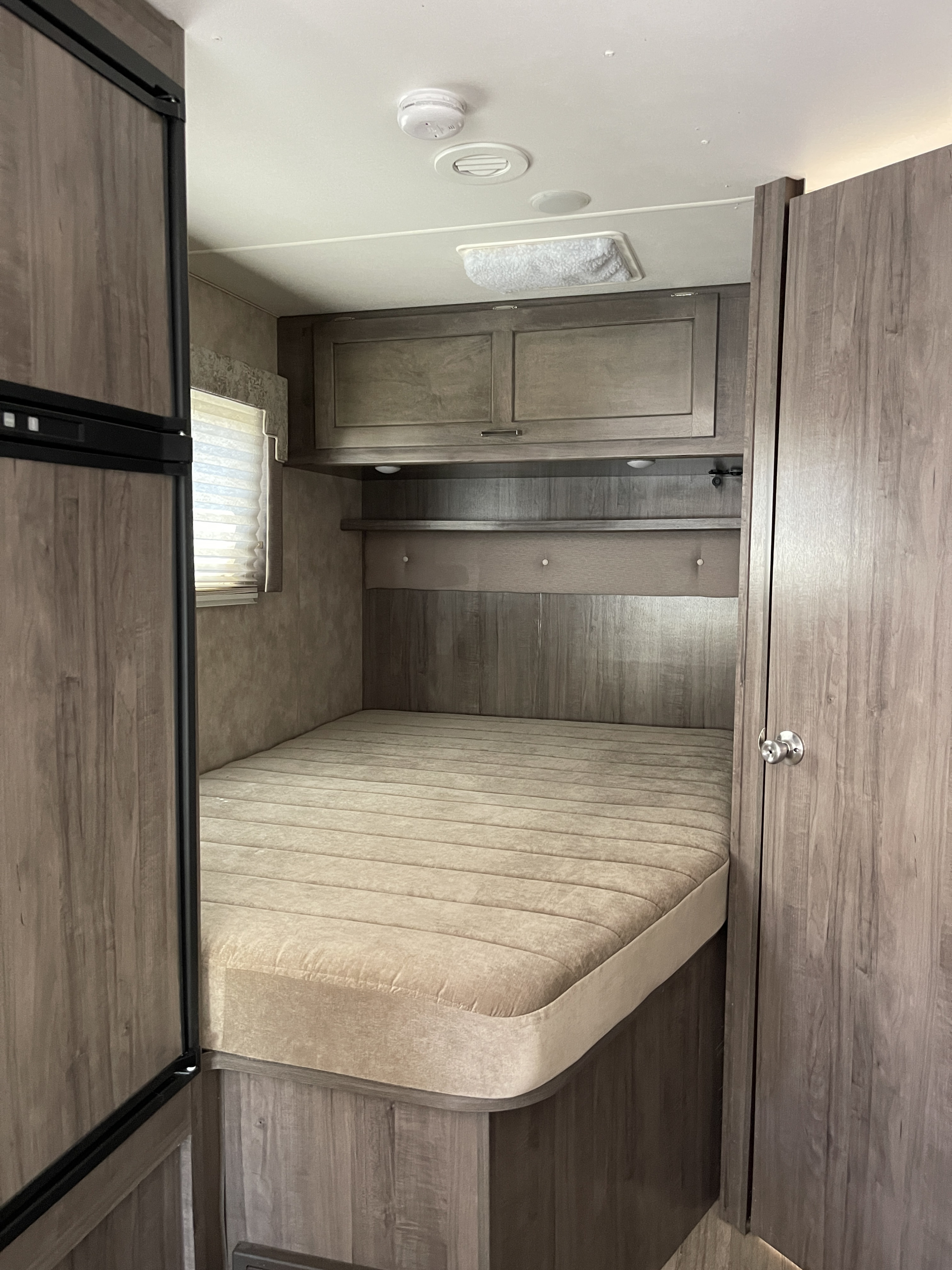 RV rental bedroom showcasing a comfortable bed, overhead cabinets, and a small window with blinds.. Winnebago Minnie Winnie 2019