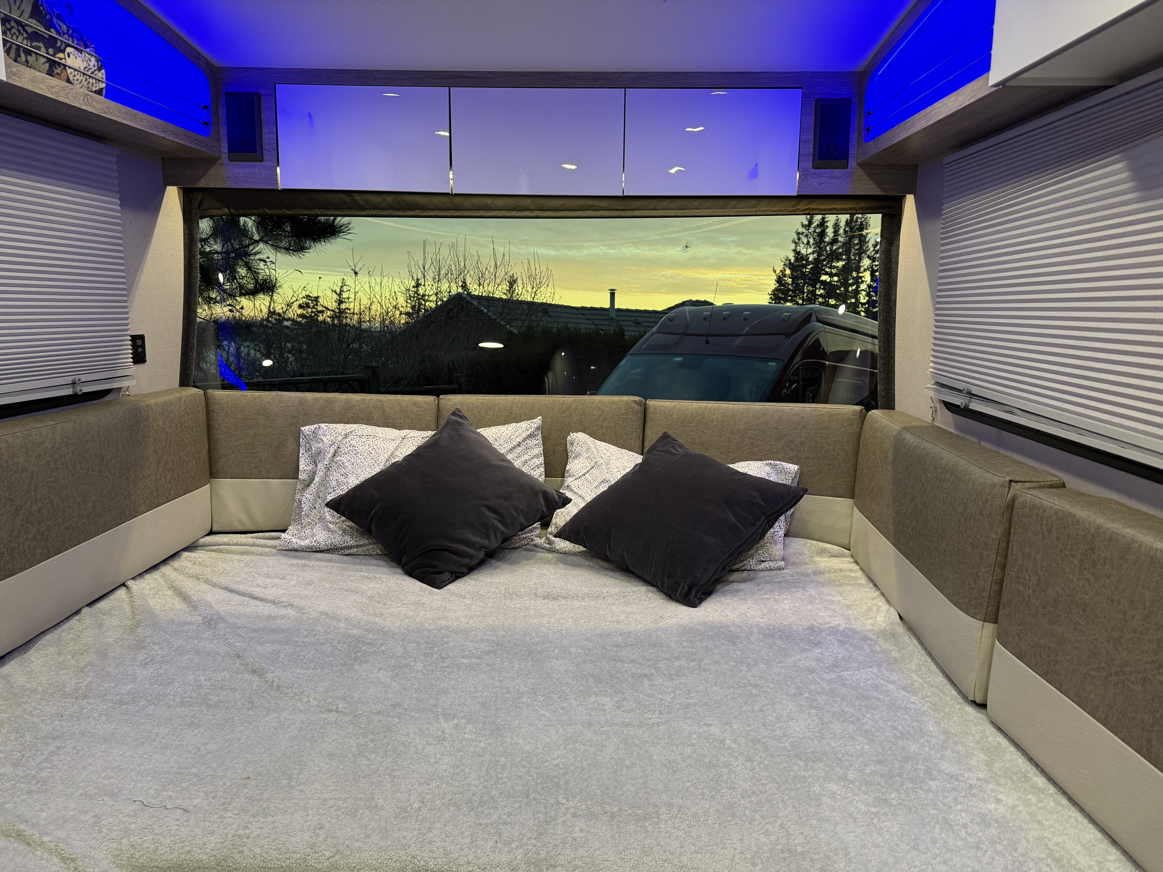 Modern RV rental interior: a comfy bed, lounge area, and window showing a beautiful sunset over a neighborhood.. InTech RV SOL 2021