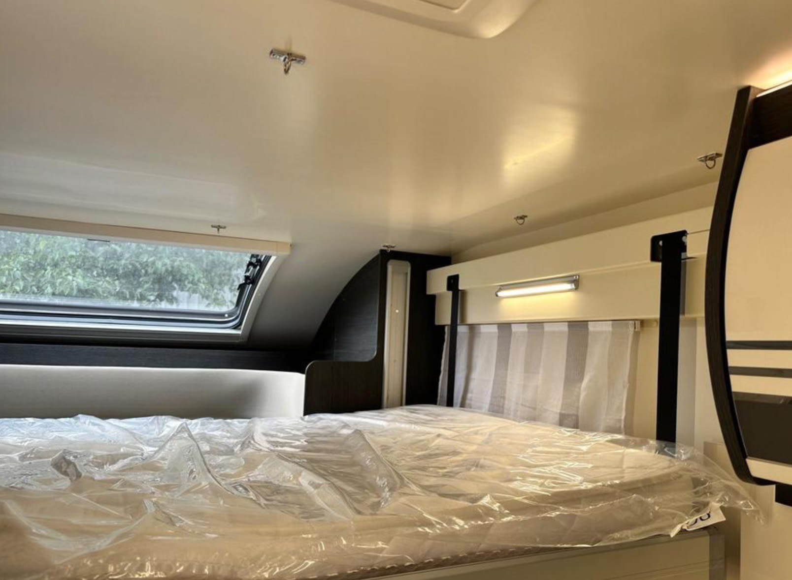 RV rental's cozy interior: bed with protective cover, large window showing trees, and integrated lighting.. Bailey Adamo 69/2 2021