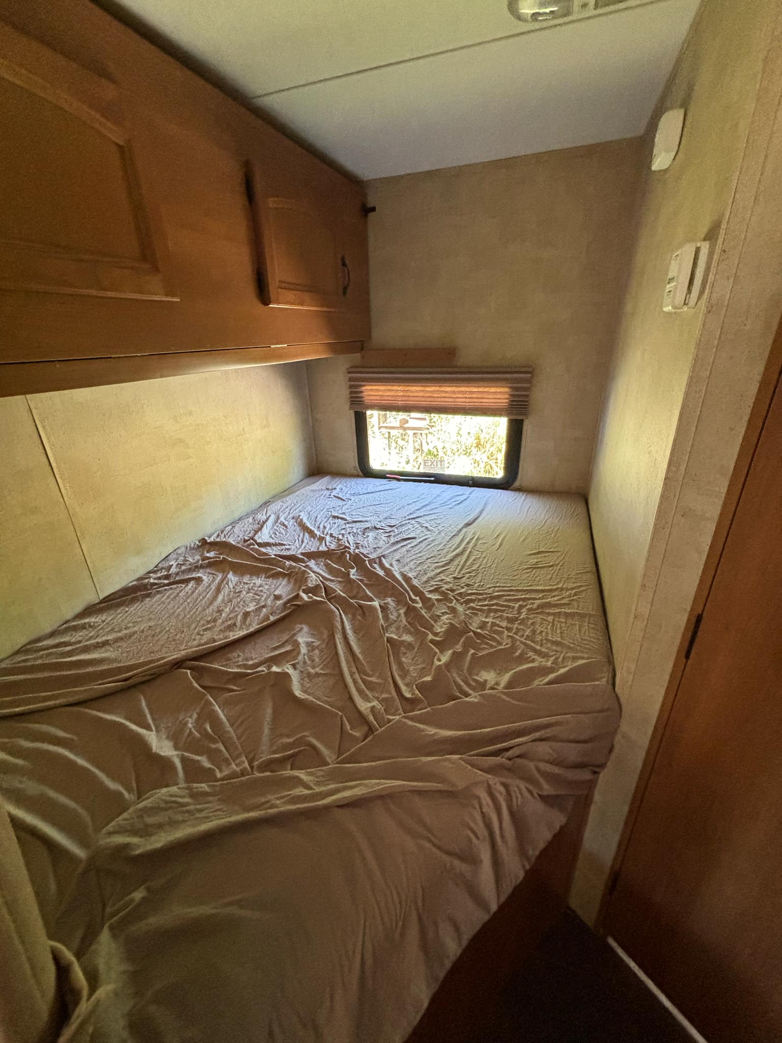Cozy RV rental bedroom features a bed with rumpled sheets, overhead cabinets, and a window with blinds.. Keystone RV Hideout 2012