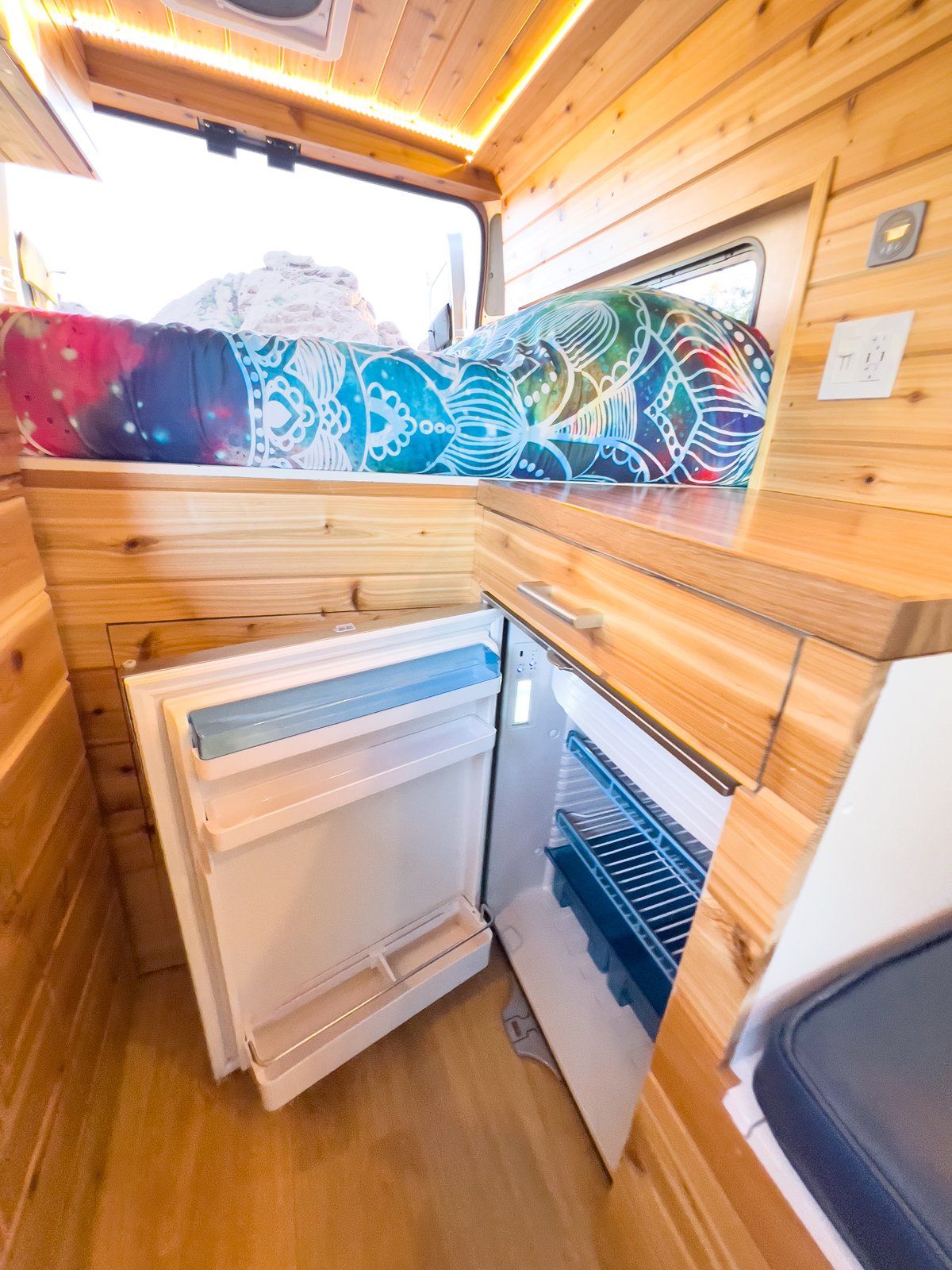 Inside an RV rental: light wood interior featuring a colorful bed, bright LED lighting, and two open mini-fridges.. Dodge RAM Promaster 2500 High Roof 2021