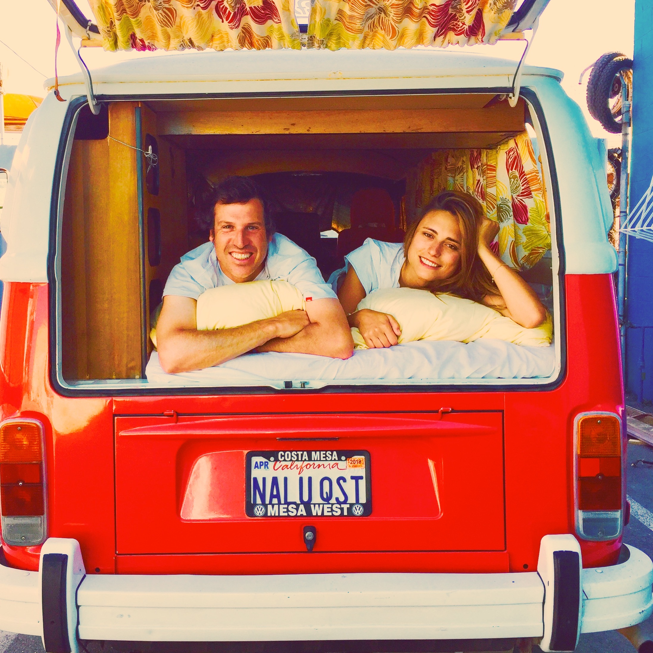 A happy couple smiles from inside a red VW campervan, set up as a cozy bed. Ideal for an RV rental getaway!. Volkswagen Westfalia 1978