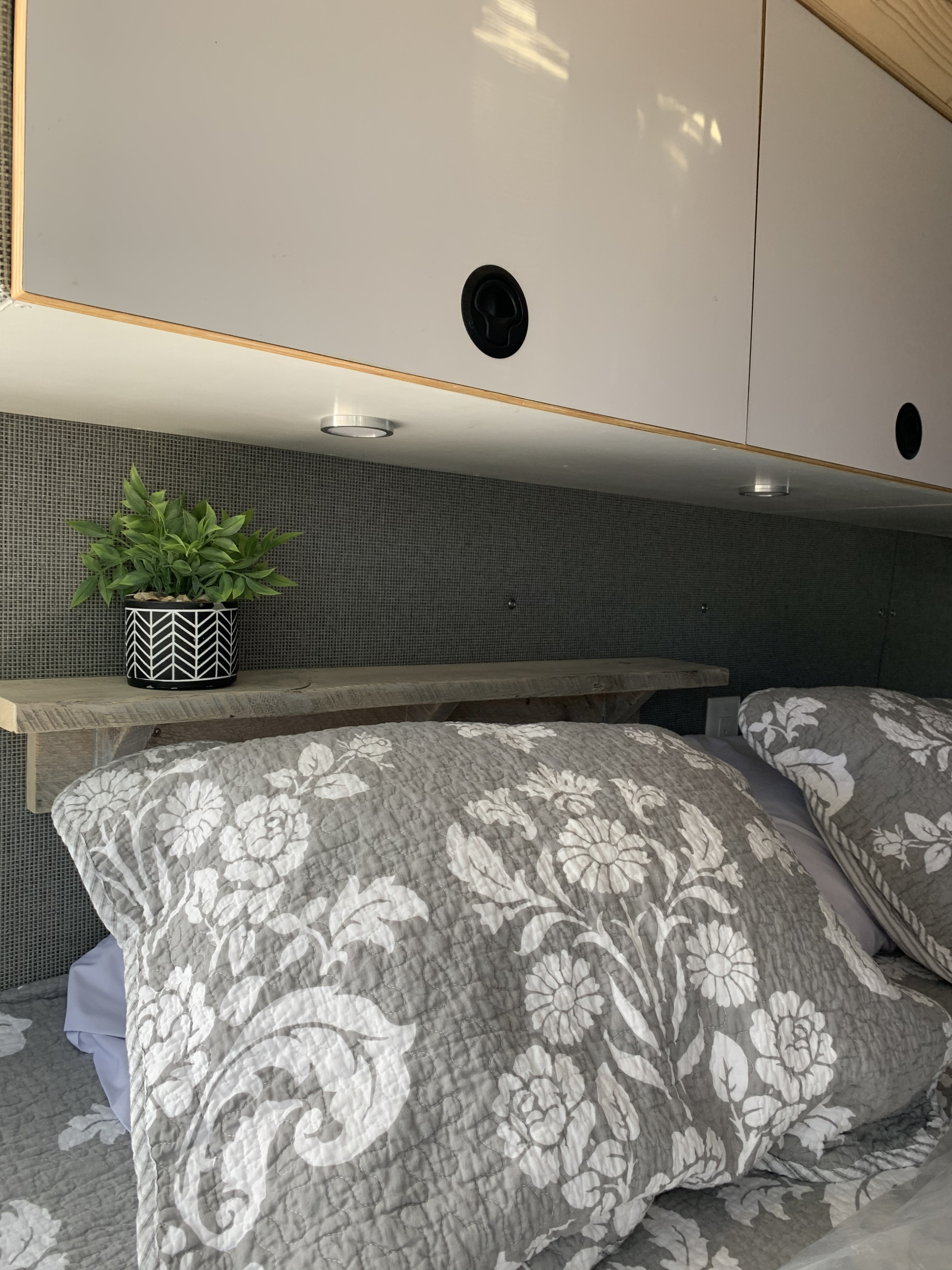 Cozy RV rental bedroom showcasing smart overhead storage, a small plant on a wood shelf, and soft floral bedding.. Ford Custom Transit 2019
