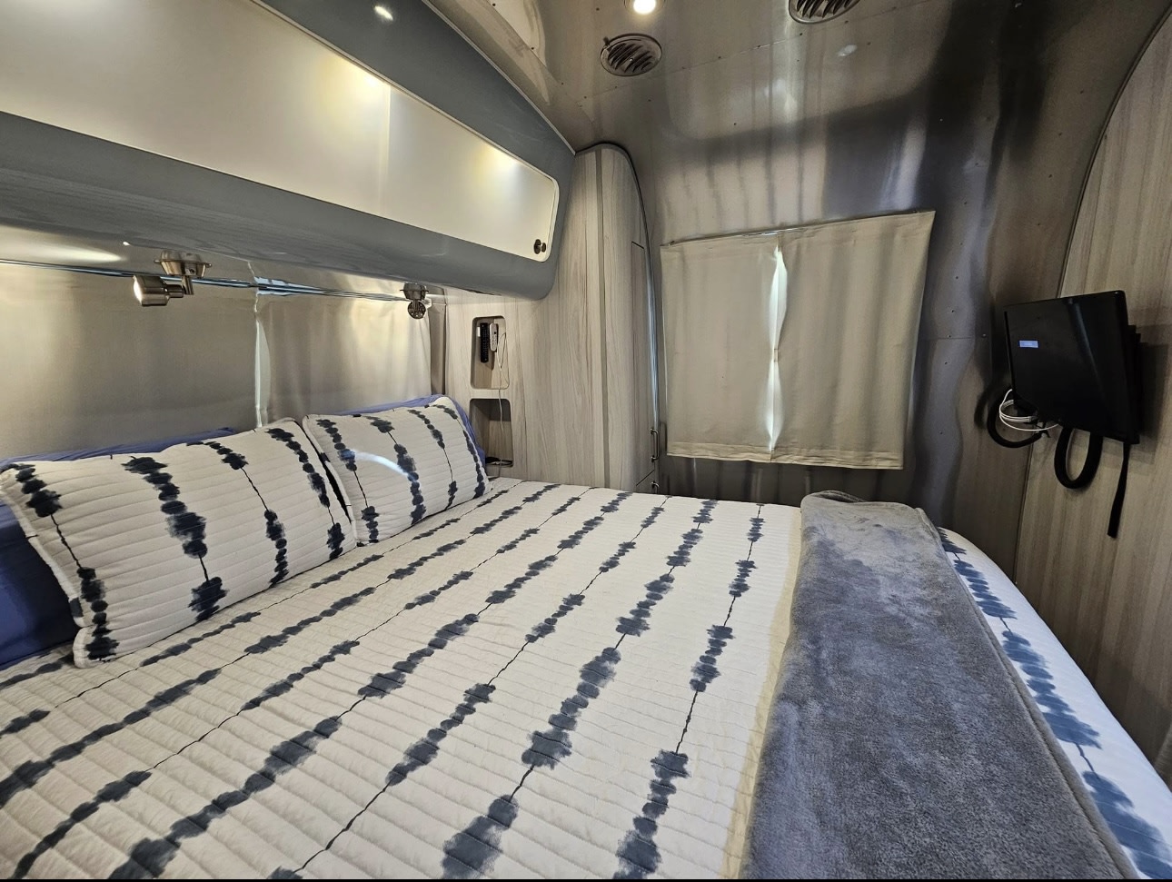 Explore this RV rental's cozy bedroom. Features striped bedding, aluminum walls, overhead lights, and a wall-mounted TV.. Airstream International 2022