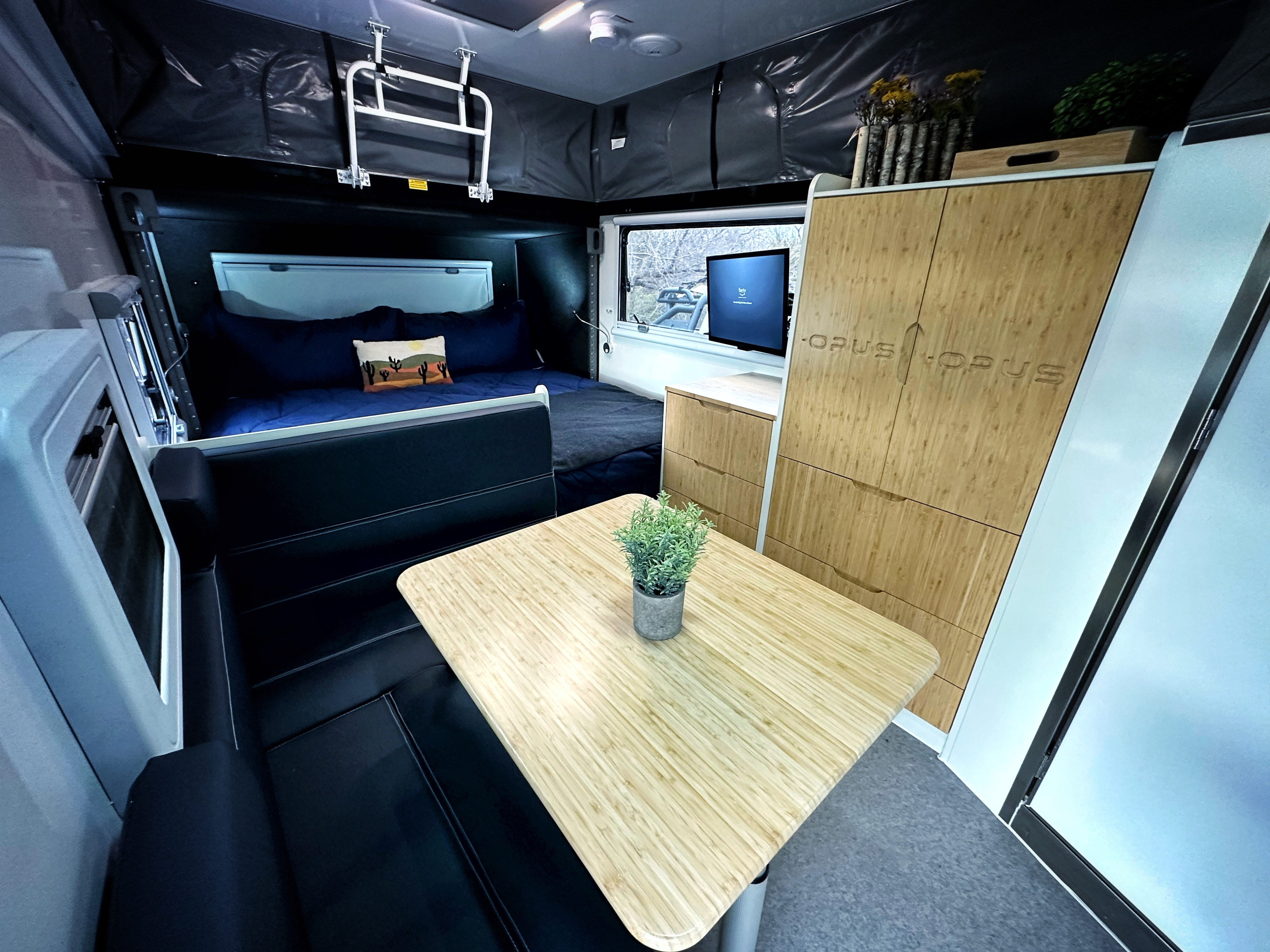 Cozy RV rental interior featuring a modern pop-up camper design with a bed, TV, wood cabinets, and dining table.. Opus Off-Road OP15 2025