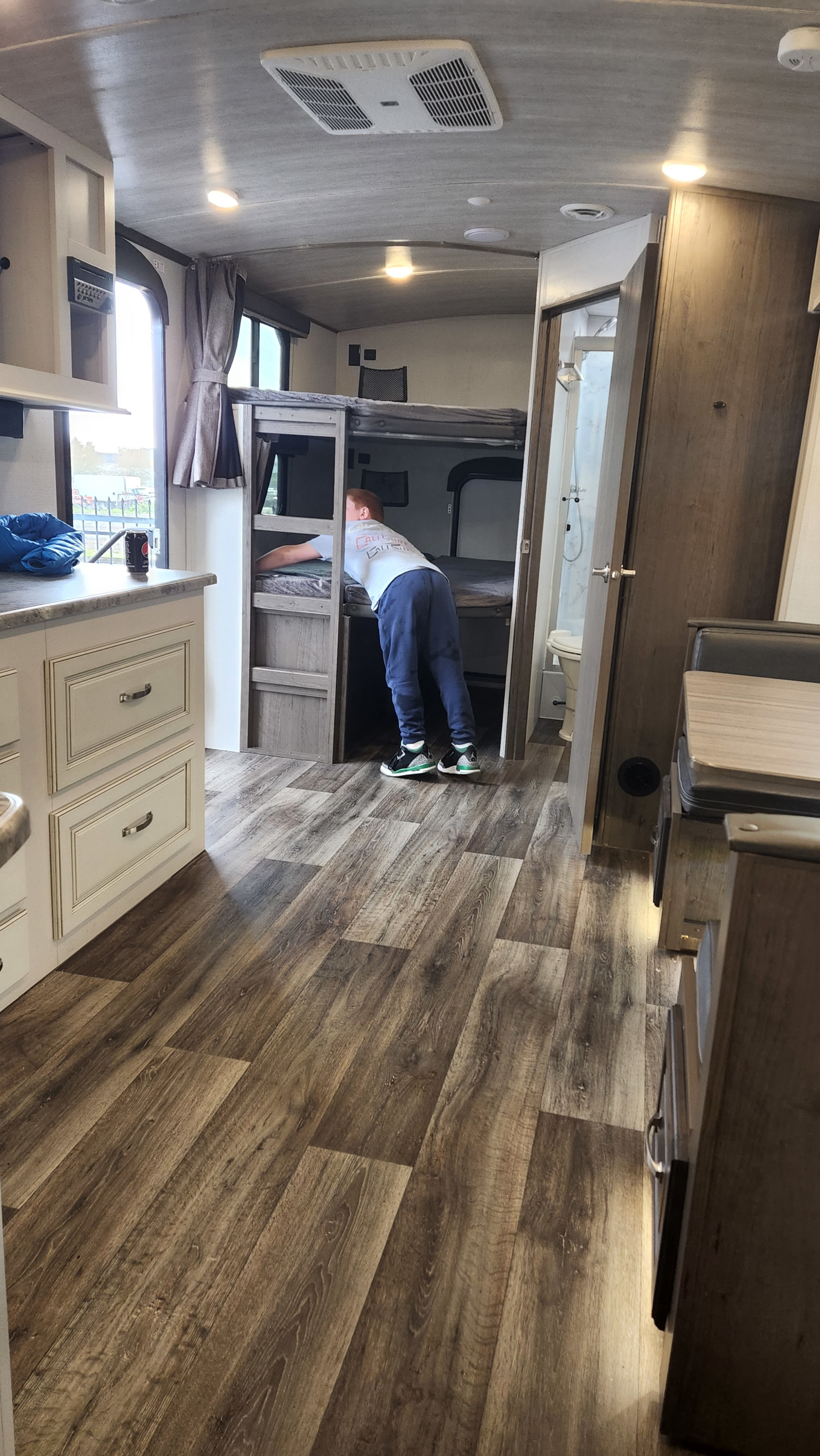 Inside an RV rental: a boy checks the bunk beds, surrounded by a functional kitchen, bath access, and wood-look flooring.. Keystone RV Cougar 2021