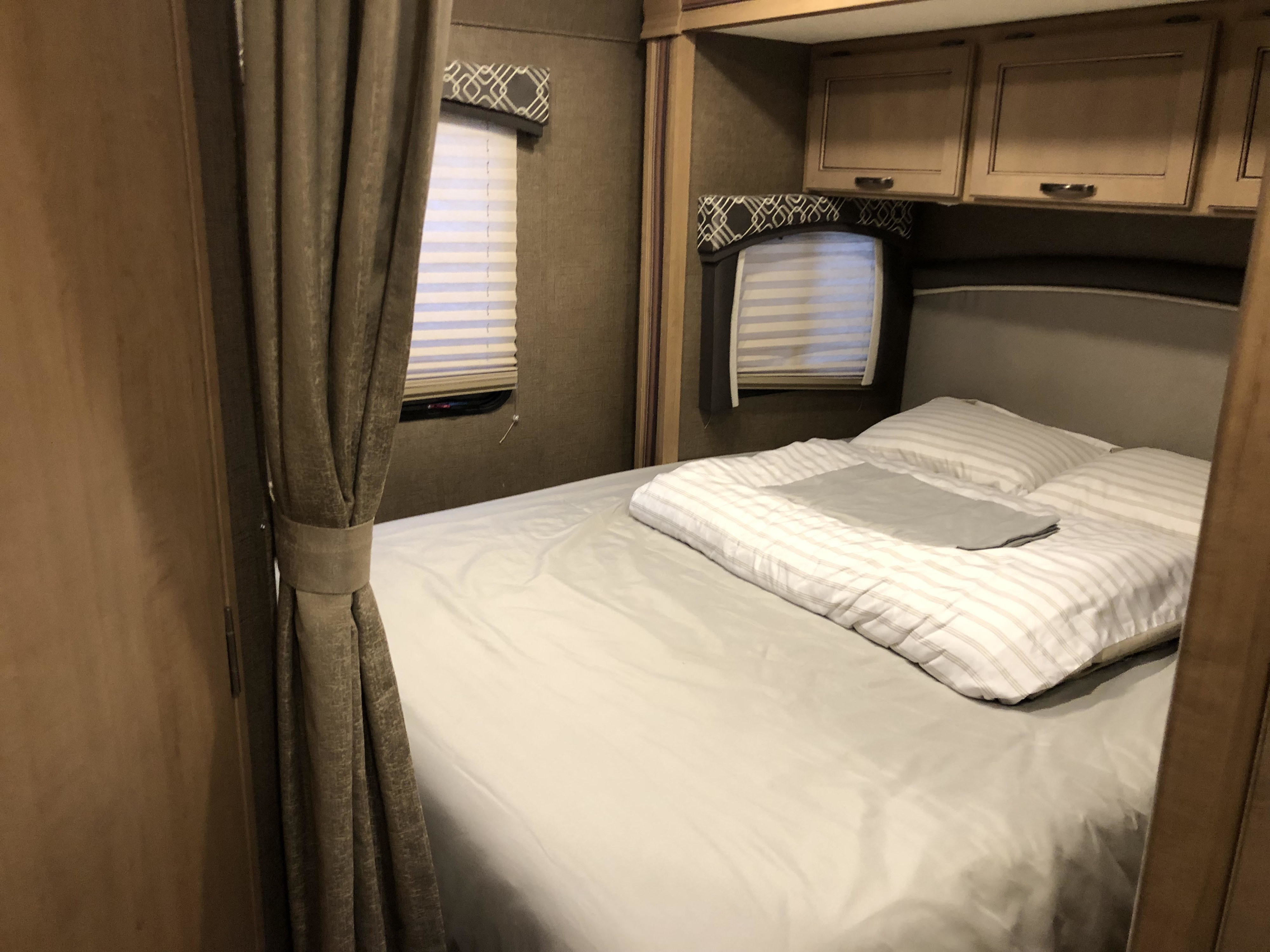 RV rental bedroom showcases a comfortable bed with striped bedding, side windows, and overhead cabinets for a cozy trip.. Thor Motor Coach Chateau 2019