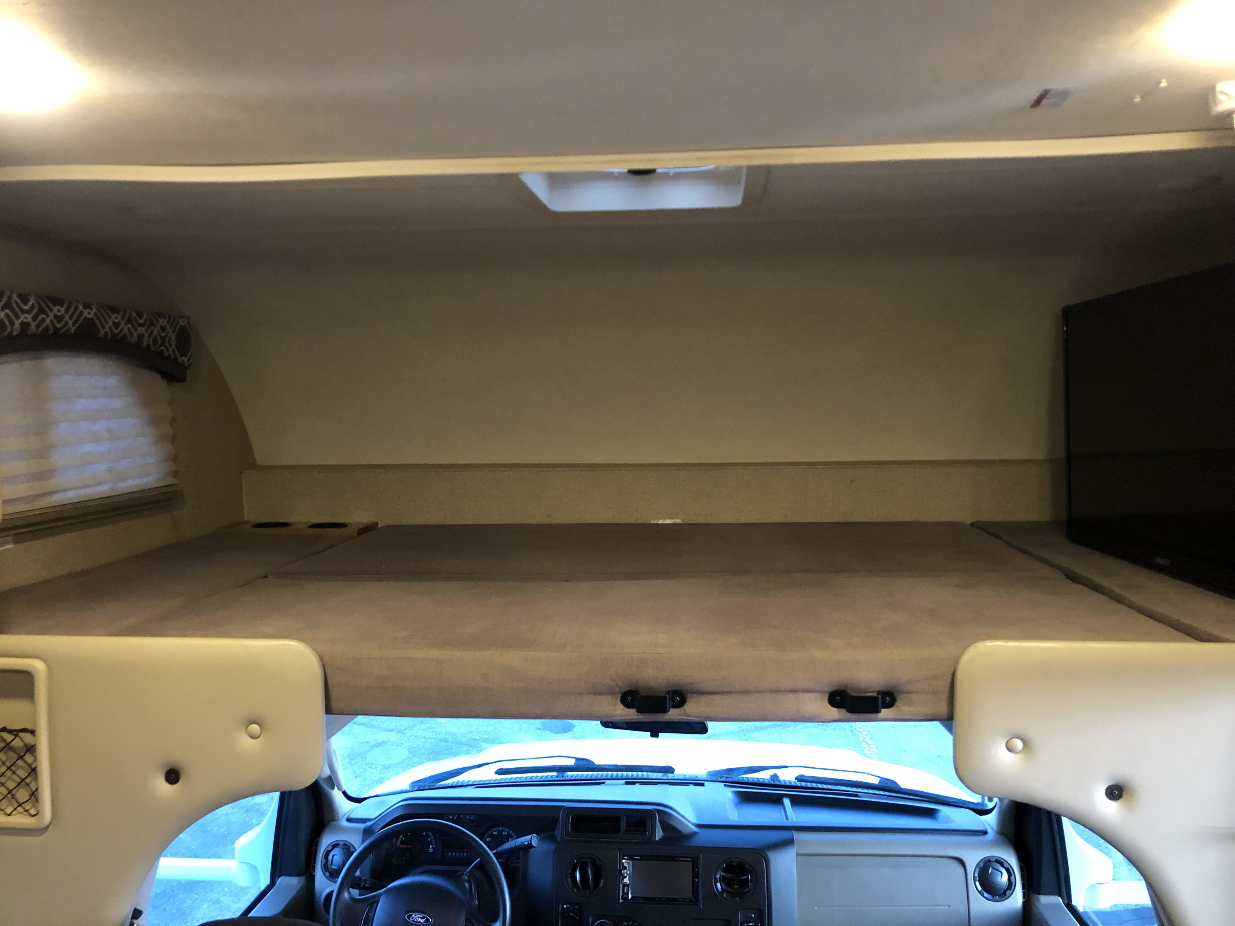 This RV rental interior showcases the cozy cab-over bed area with a window, TV, and the vehicle's dashboard and cockpit below.. Thor Motor Coach Chateau 2019