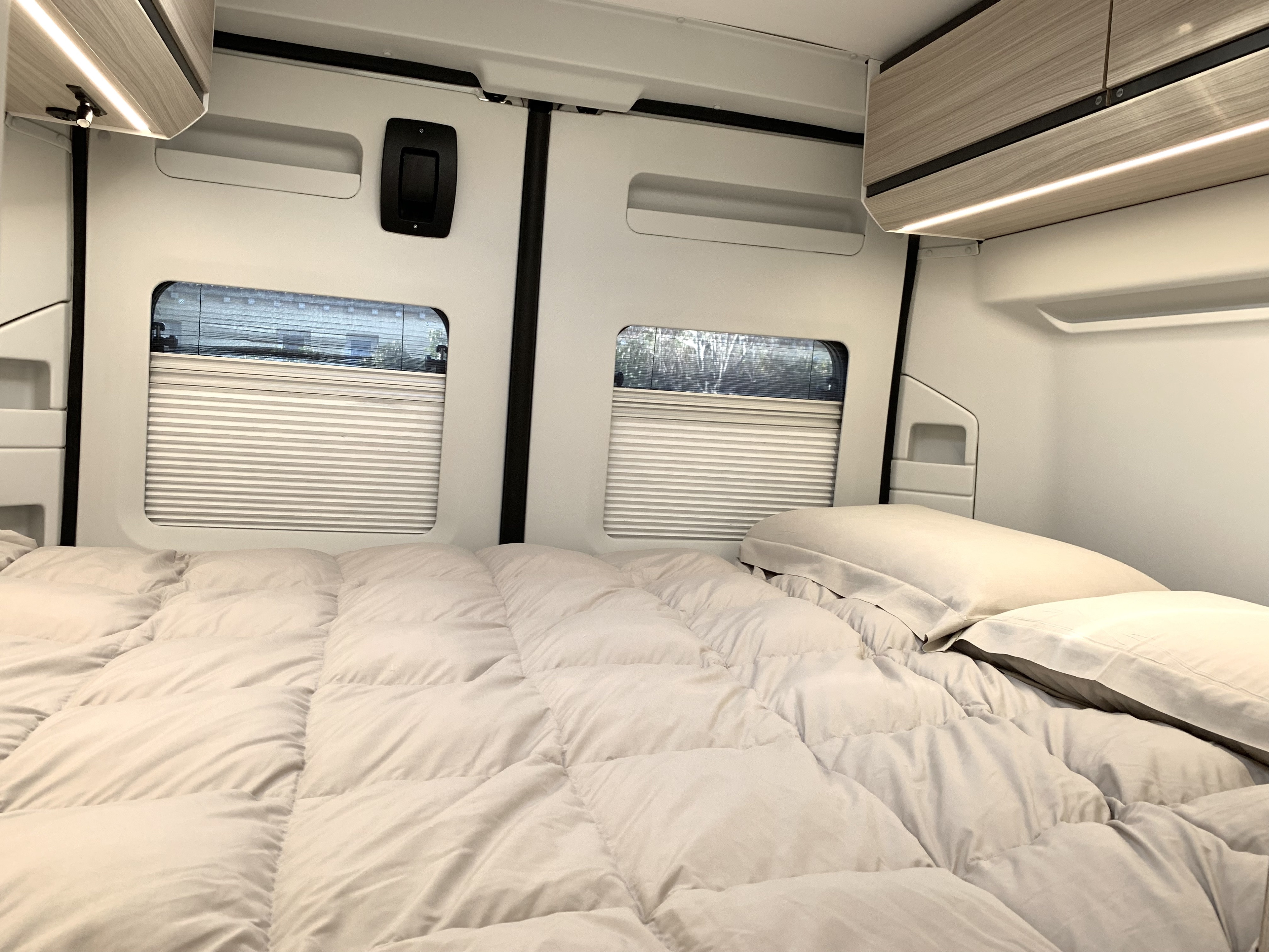 Inside a clean RV rental: a modern bedroom with a comfortable bed, two windows, and lighted overhead cabinets.. Adria Twin 600 SP 2024