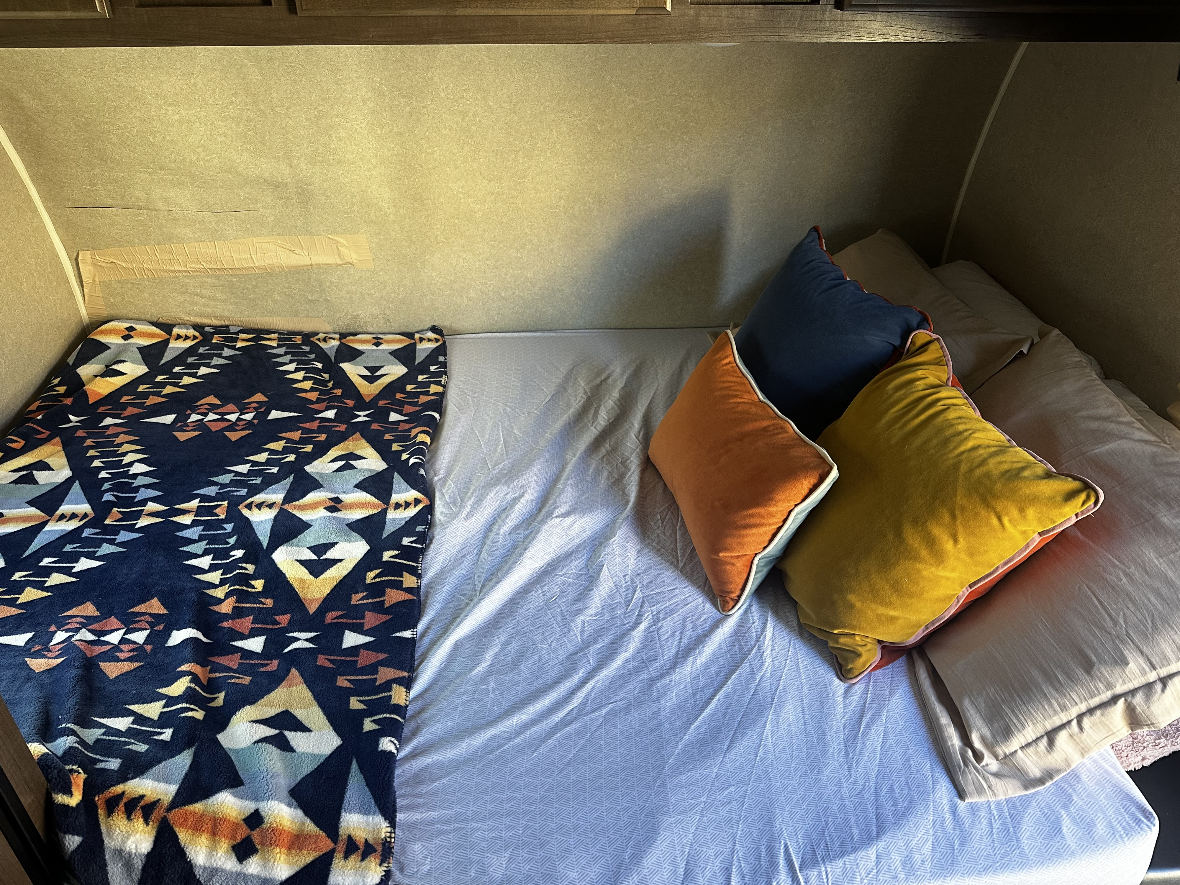 An RV rental bed featuring a blue patterned blanket, grey sheet, and stacked pillows in navy, orange, and yellow. Cozy camper interior.. Sunset Park RV Sun Lite 2020