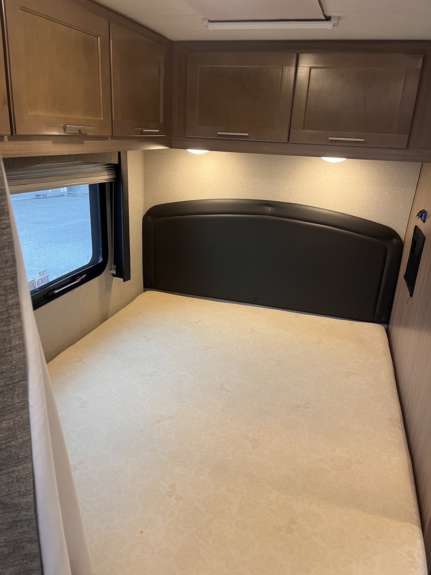 RV rental bedroom featuring a bed with black headboard, wood overhead cabinets, and window. Comfortable travel.. Ford Adventurer 2021