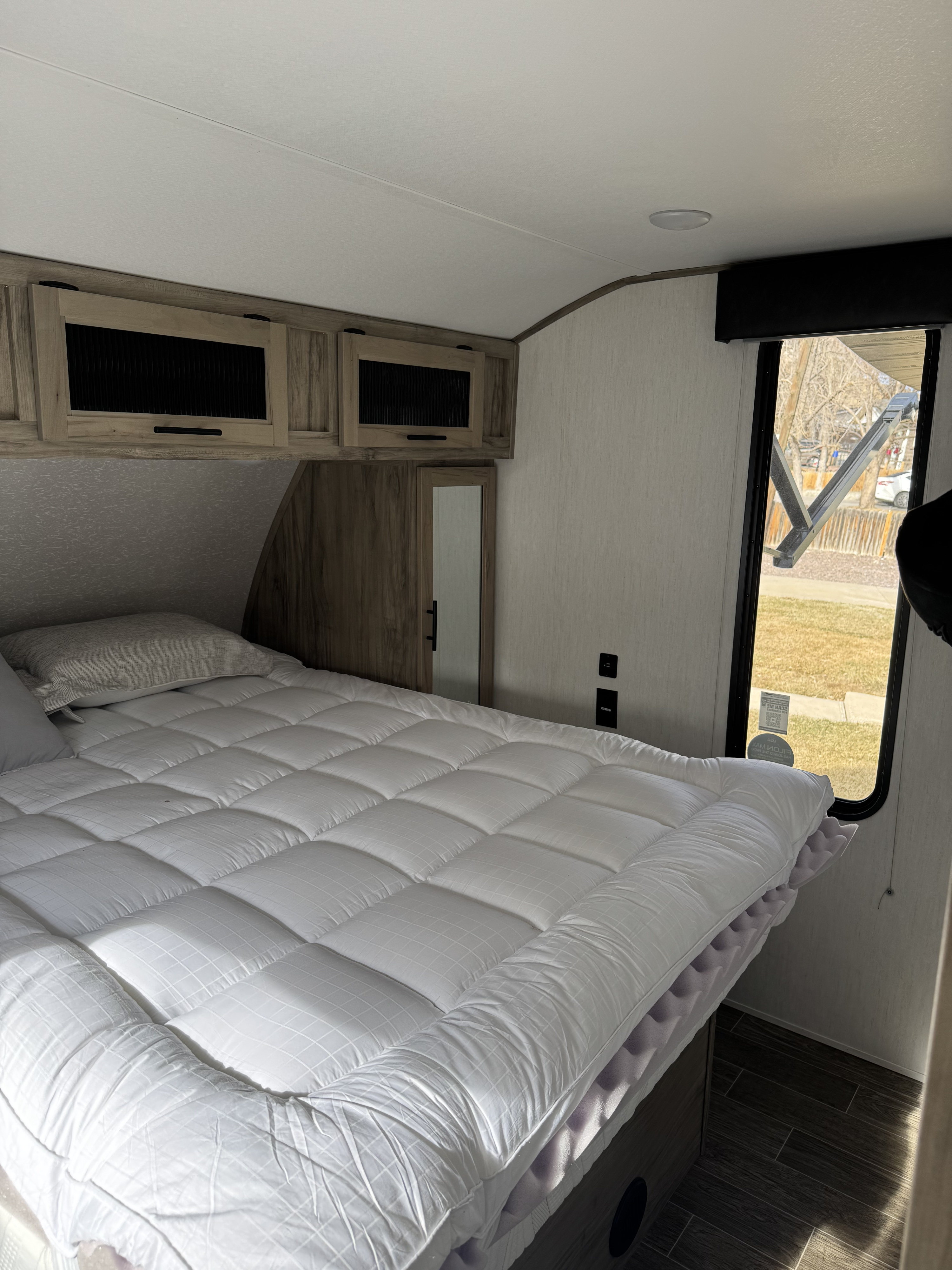 Cozy RV rental bedroom featuring a white bed, overhead wooden cabinets, and a window offering an outdoor view.. Forest River Cherokee Alpha Wolf 2022