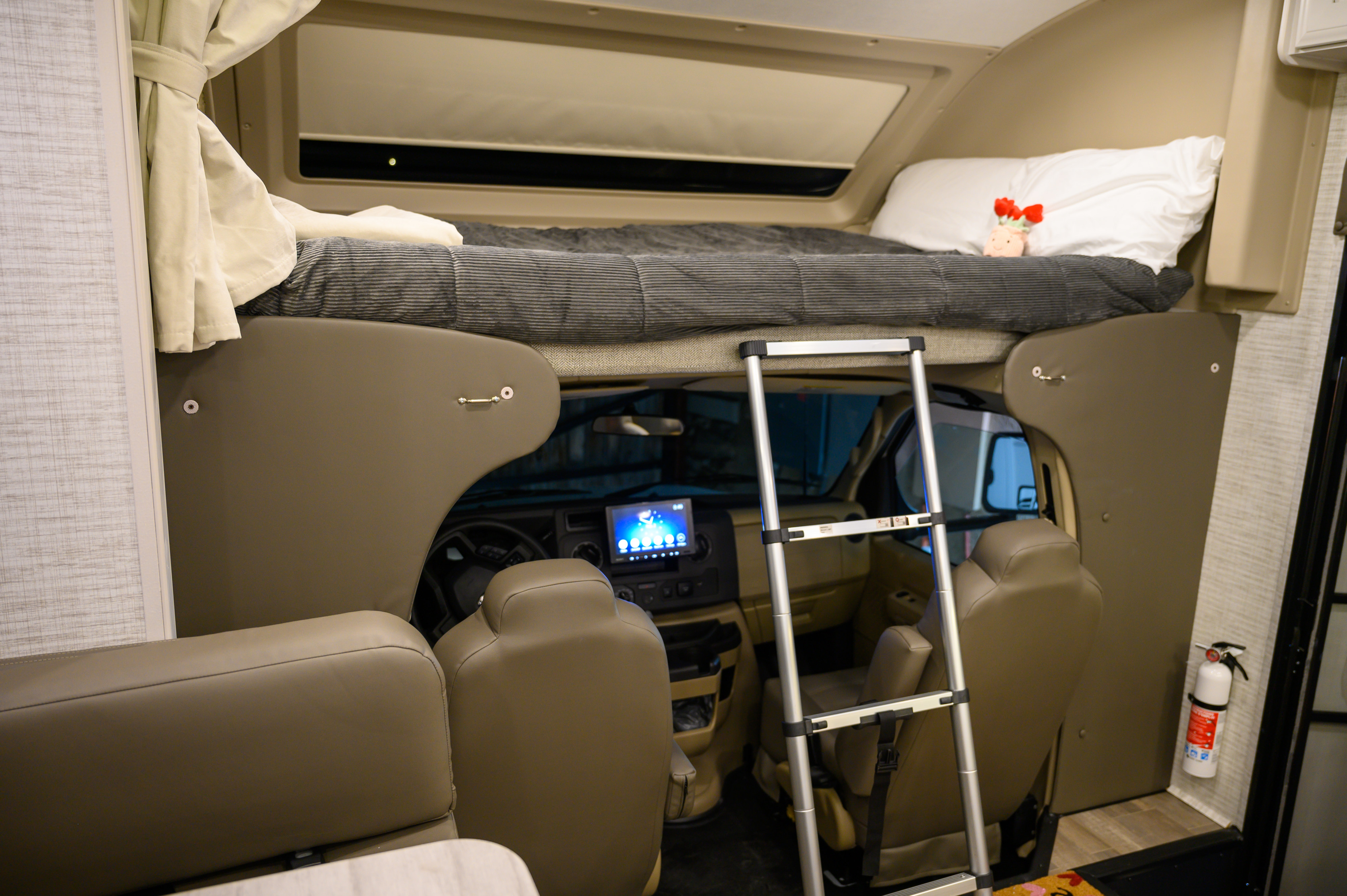 Cozy RV rental interior showing a cab-over bed with ladder, overlooking the cockpit and seating area. Features a fire extinguisher.. Entegra Odyssey 2025