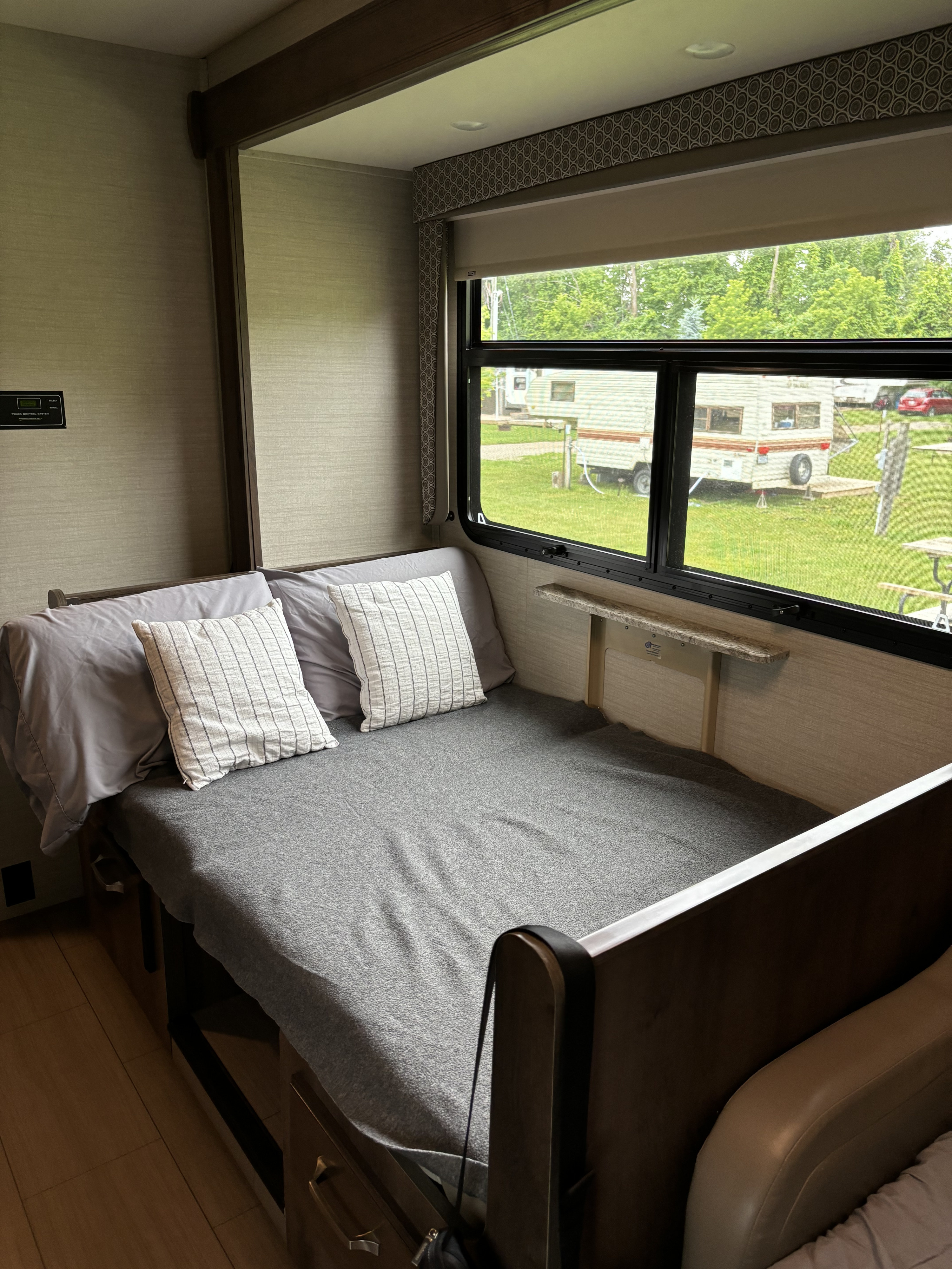 RV rental interior featuring a cozy bed with grey bedding and white pillows, beside a large window overlooking a lush green campground.. Jayco Greyhawk 2018