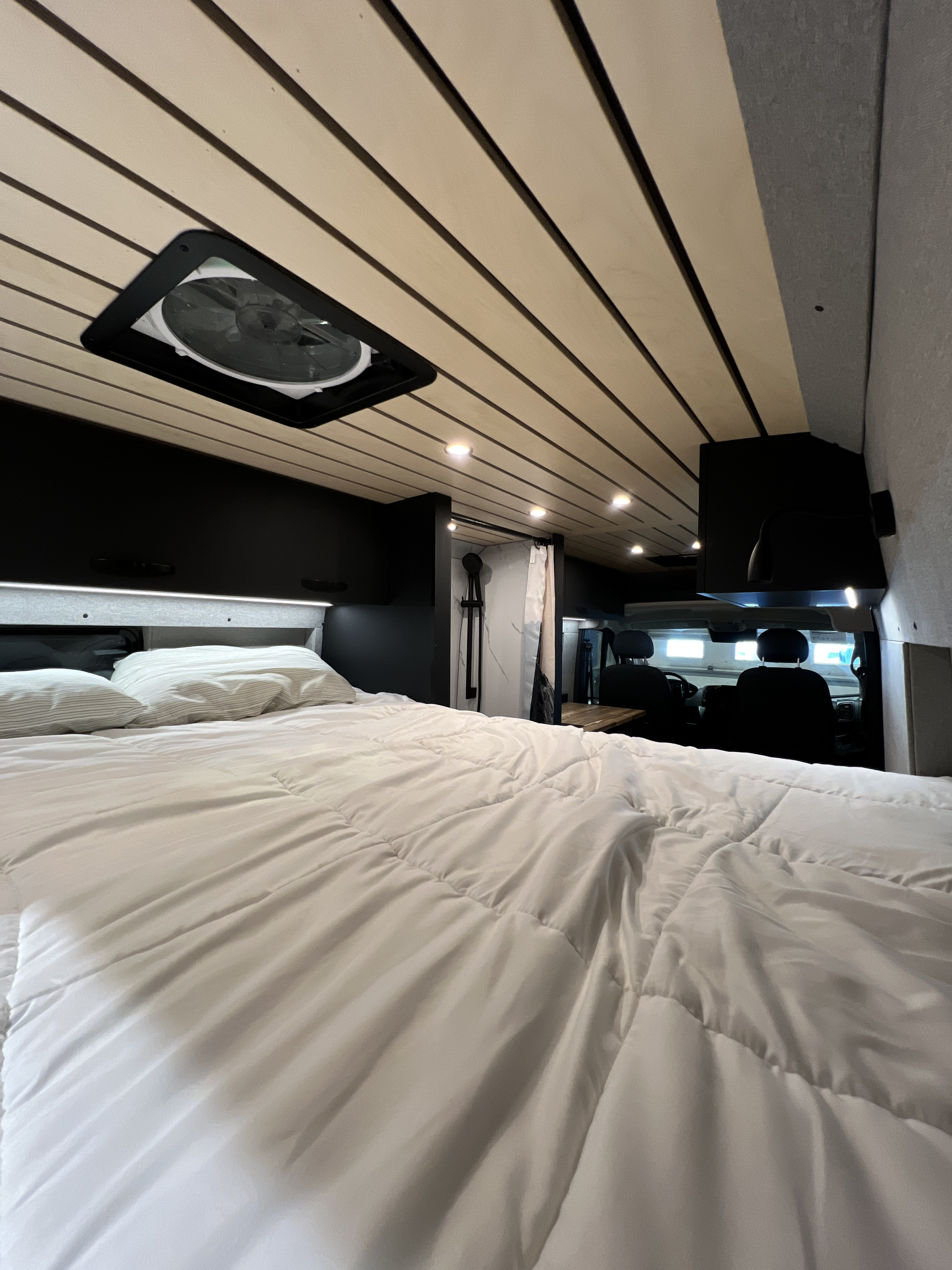 Spacious RV rental conversion showcasing a comfortable bed, wood-slat ceiling with fan, and compact shower area.. Dodge Ram Promaster 2025