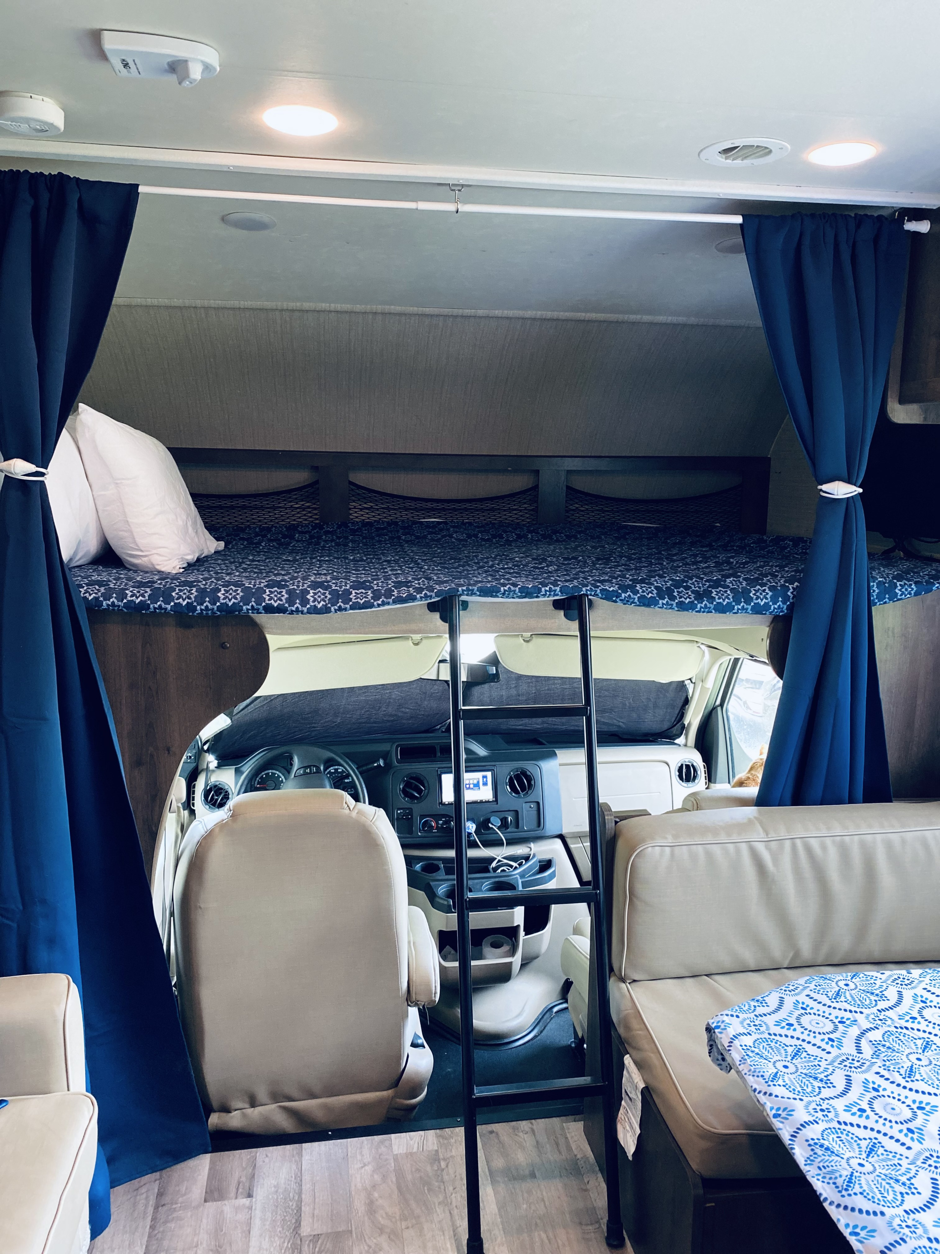 Inside an RV rental: a cozy overhead bunk with blue bedding and ladder, driver's cab, and seating area with curtains.. Jayco Redhawk 2019