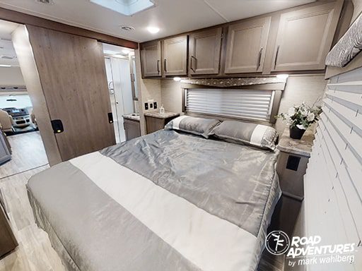 Experience the comfortable RV rental bedroom, featuring modern grey bedding, overhead cabinets, and a sliding door to the living space.. Thor Motor Coach Four Winds 2023