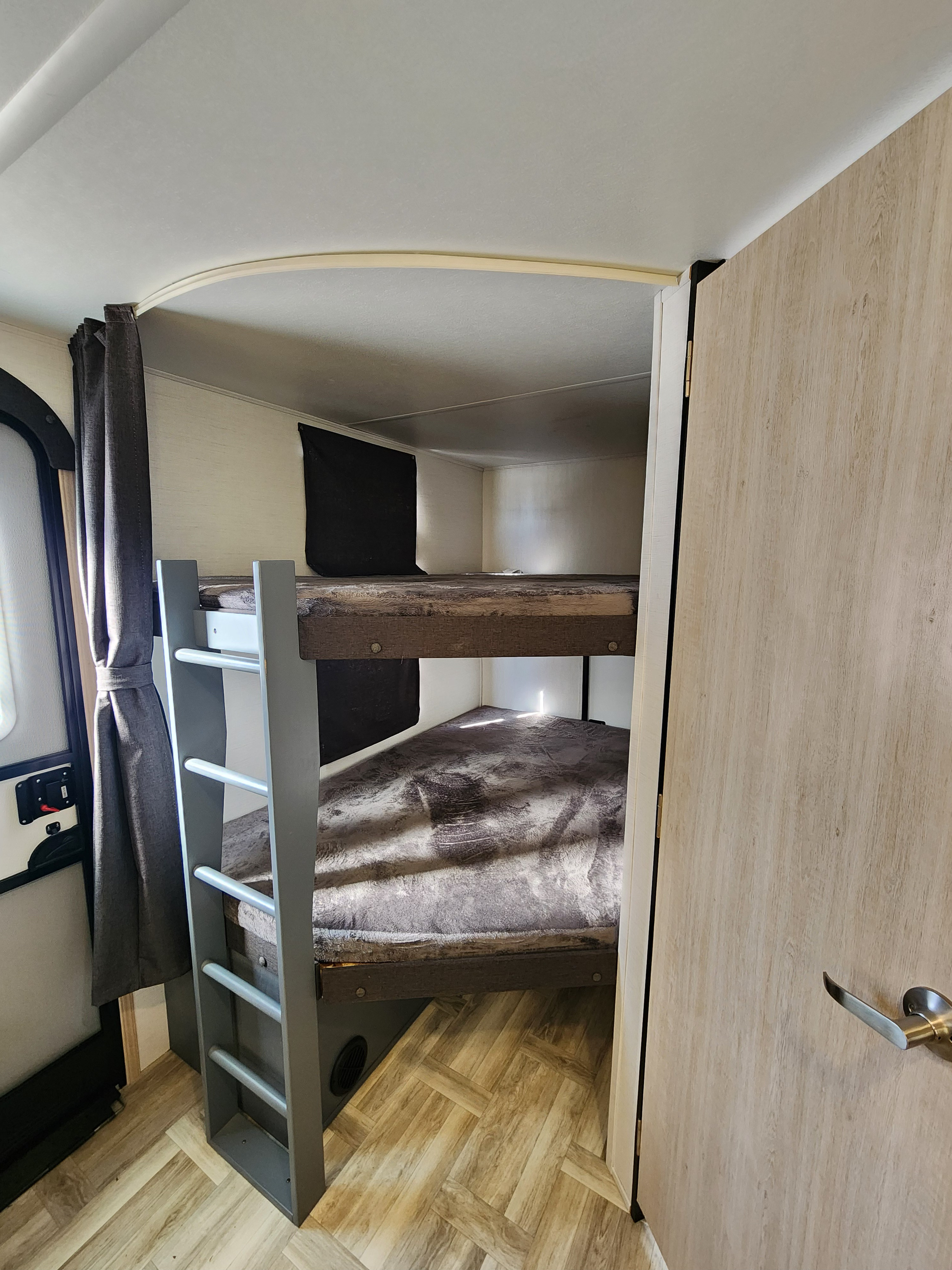 RV rental bunk beds: a compact sleeping area with a gray ladder, light wood-look floor, and window curtain.. primetime tracer 2024