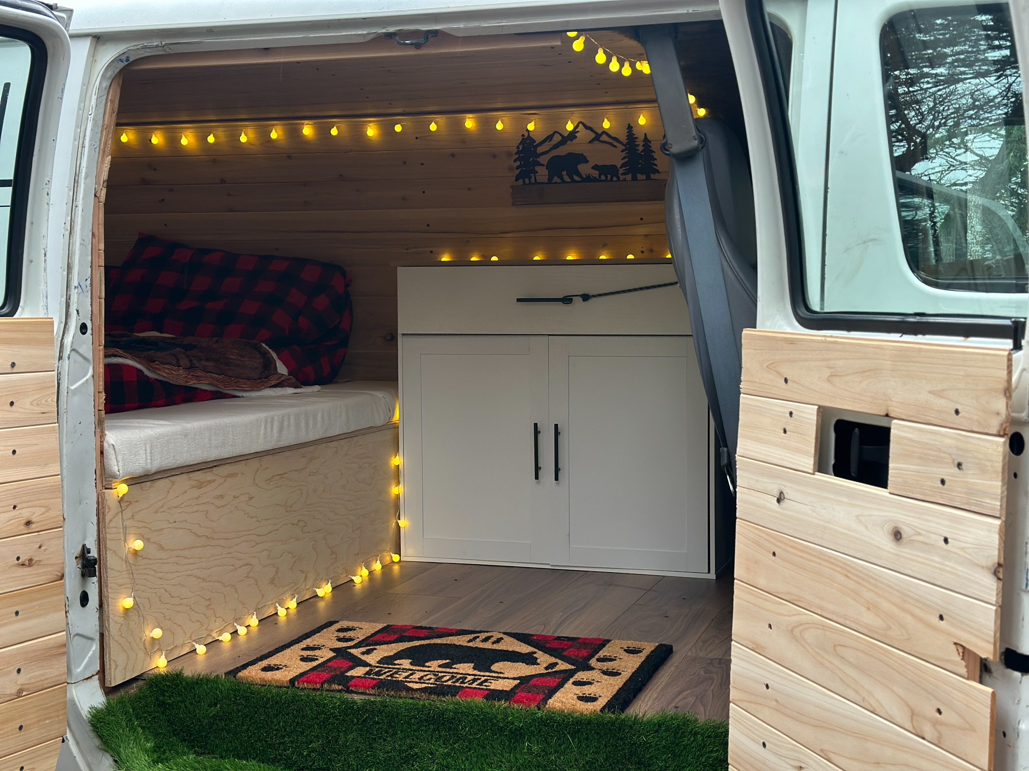 Cozy rv rental camper van interior. Features wood walls, a bed with plaid blanket, string lights, cabinets, and bear decor.. Dodge Ram Van Roadtrek 2000