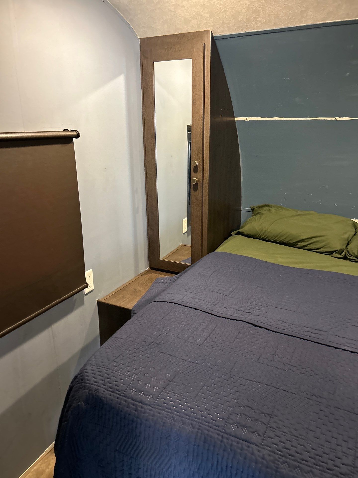 Compact RV rental bedroom with a blue quilted bed, olive sheets, and a built-in mirrored cabinet for convenience.. Forest River Wildwood 2019