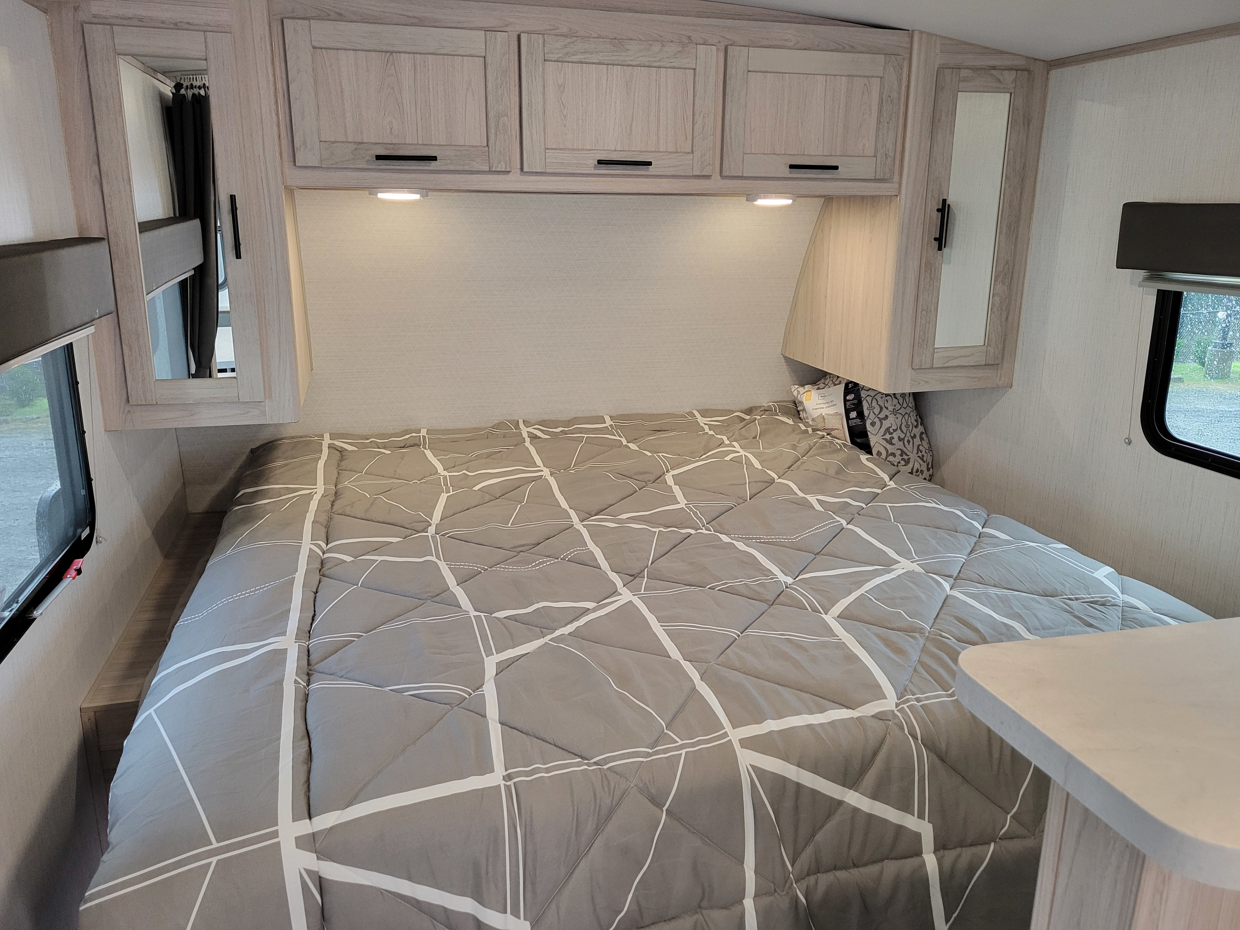 Cozy RV rental bedroom featuring light wood cabinets, under-cabinet lighting, and a gray patterned comforter on the bed.. Cruiser RV Shadow Cruiser 2022