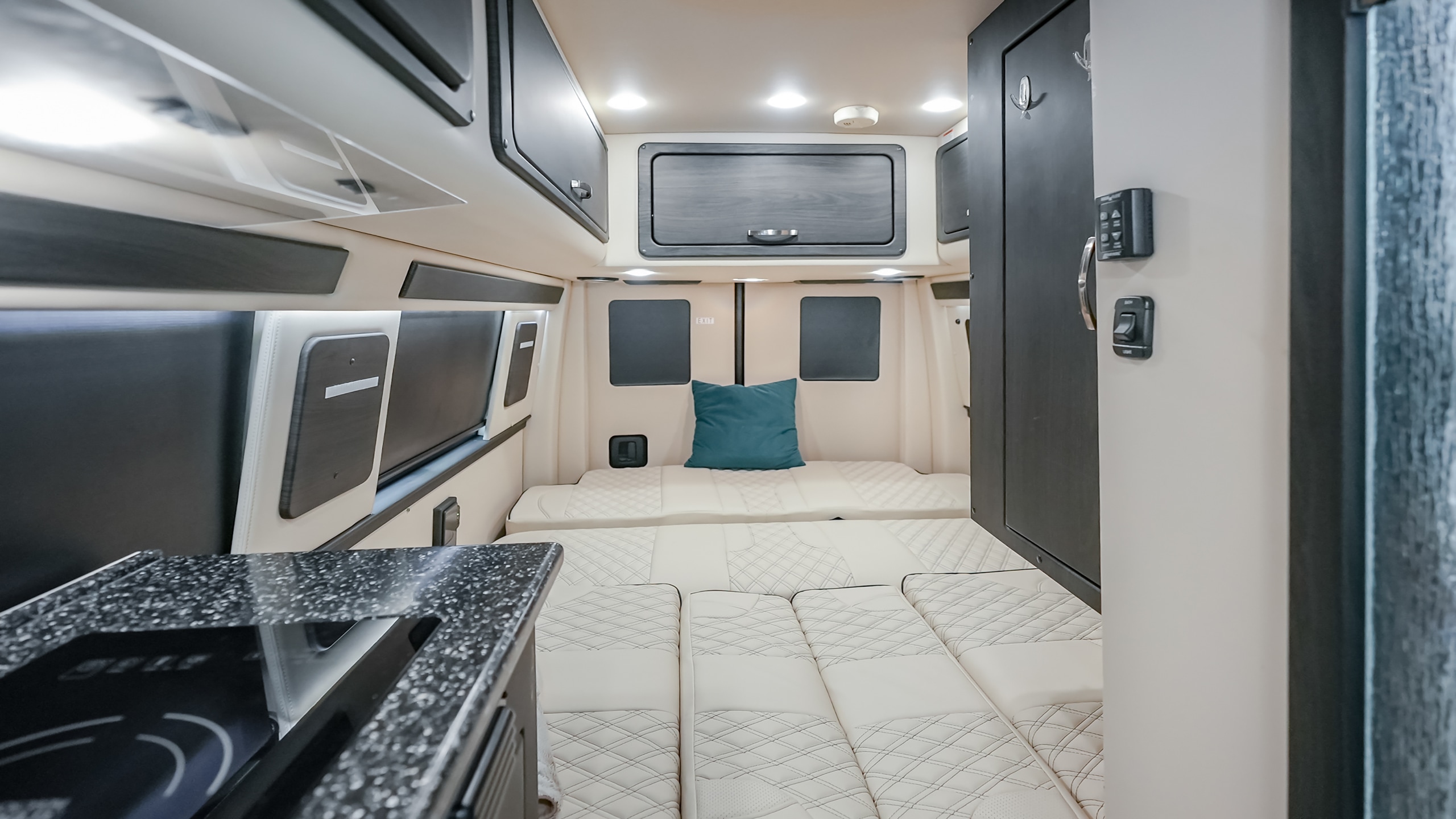 RV rental van interior showcasing modern dark cabinetry, cozy cream quilted bed/seating, and a compact kitchenette.. American Coach Mercedes-Benz Sprinter 2023