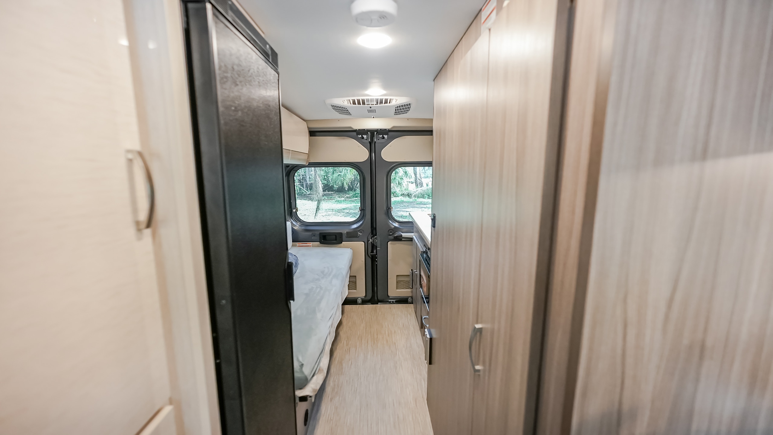 RV rental interior: Hallway view of a cozy camper van with a bed, refrigerator, and light wood cabinets.. Jayco swift 2024