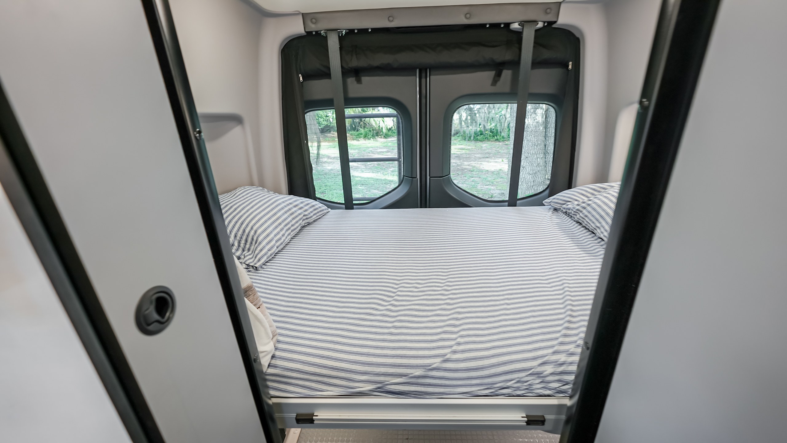 Neatly made bed in an rv rental, with striped sheets and green outdoor scenery visible through the rear windows.. Winnebago Mercedes-Benz 2023