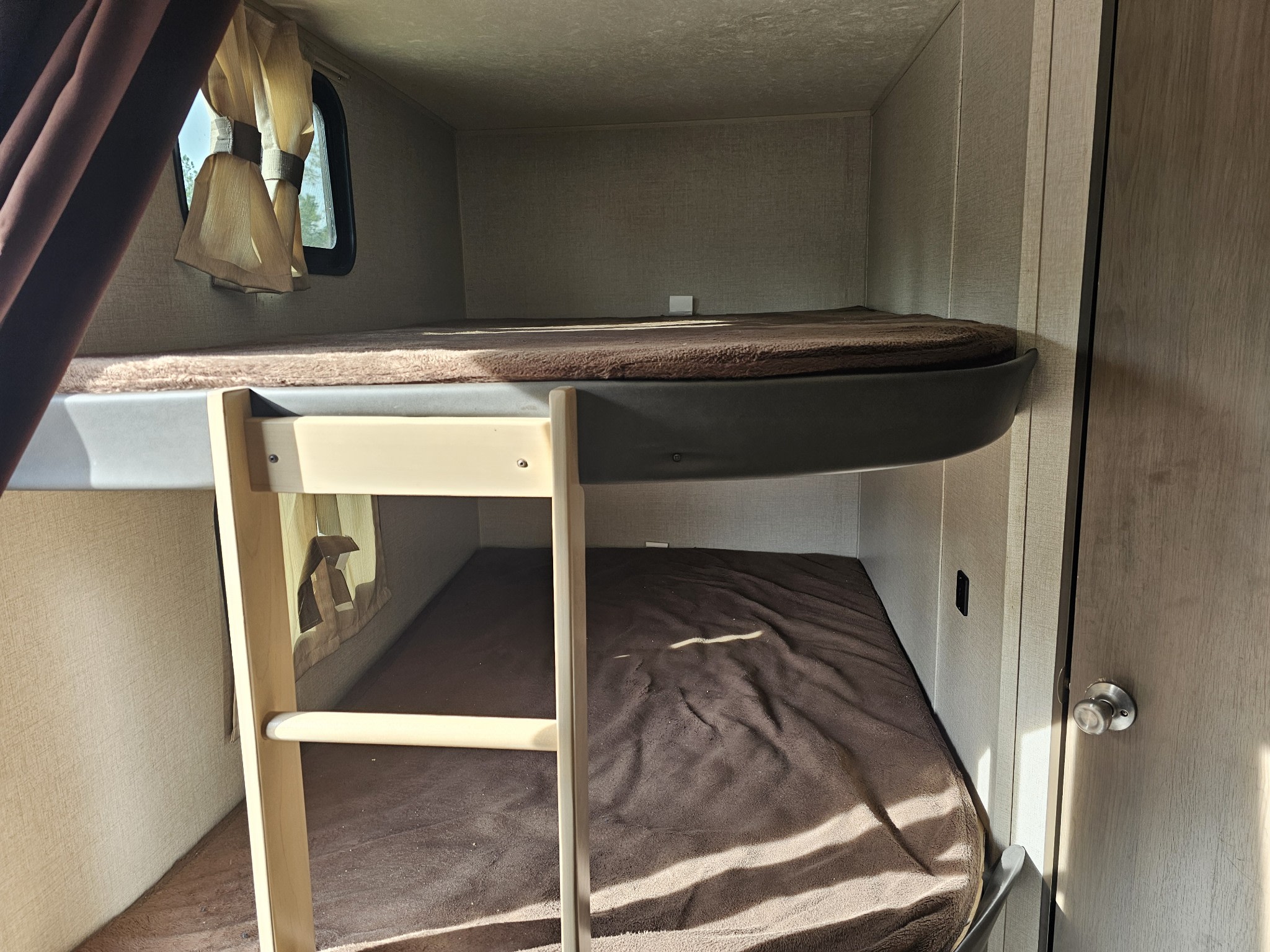 This RV rental features cozy bunk beds with brown blankets and a wooden ladder. A small window with beige curtains provides natural light.. Forest River Catalina 2020