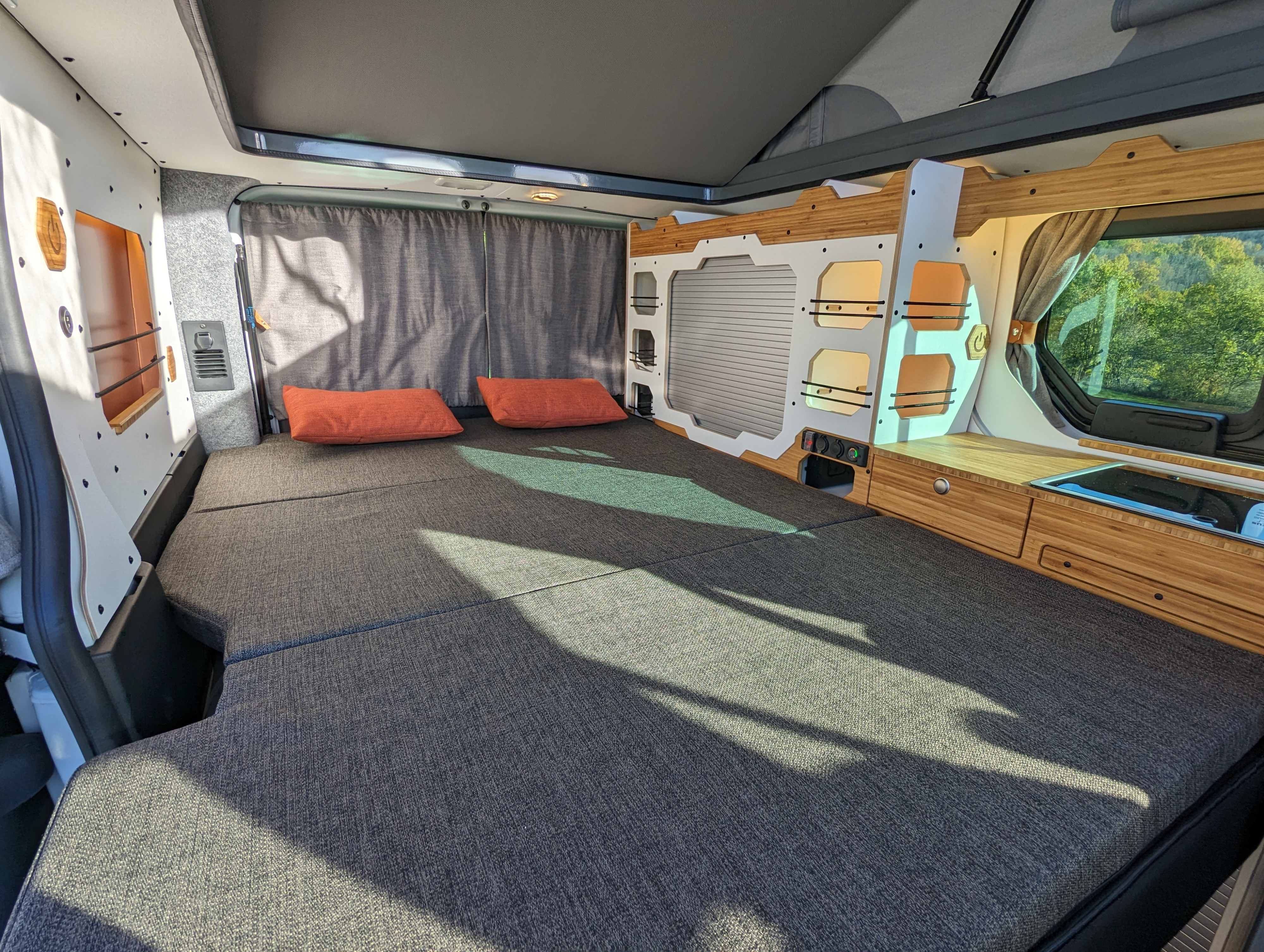 Cozy RV rental camper van interior. Features a large bed with orange pillows, bamboo storage, and a built-in sink.. Renault Trafic 2025