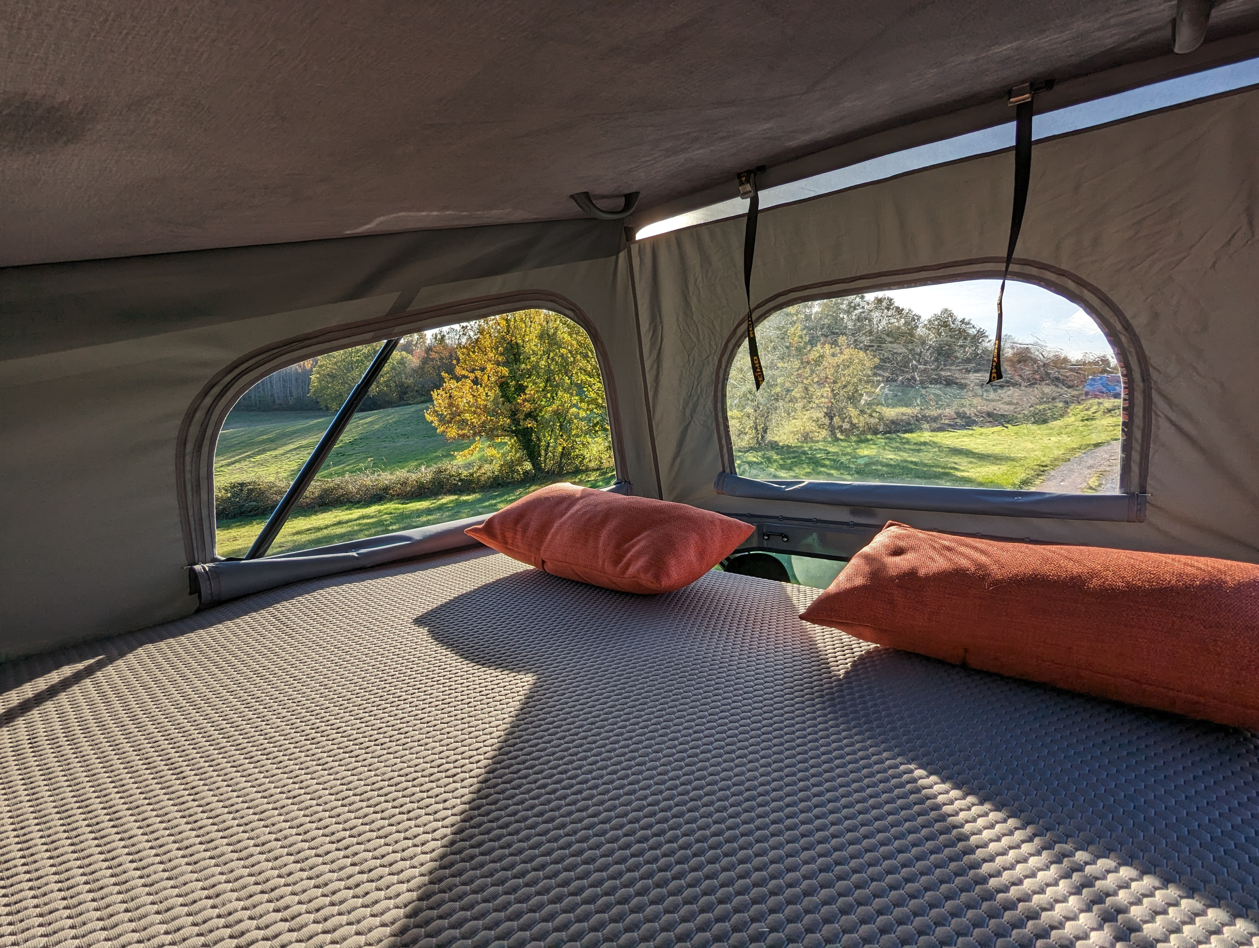 Cozy pop-top RV rental bed with orange pillows, offering a bright, scenic view of green fields and trees through large windows.. Renault Trafic 2025