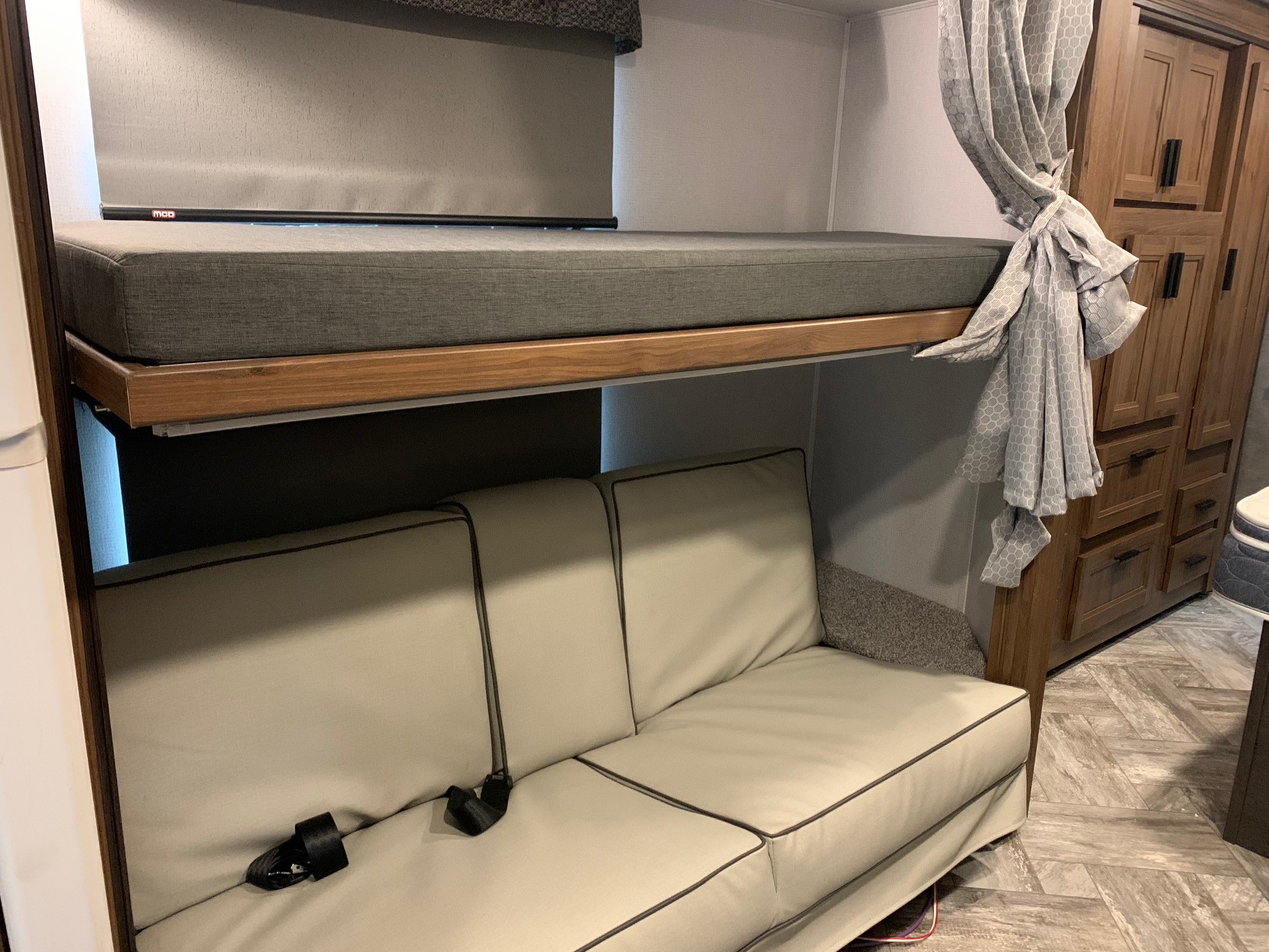 RV rental interior: a grey bunk bed over a light sofa with seatbelts, next to wood cabinets.. Forest River Sunseeker 2026