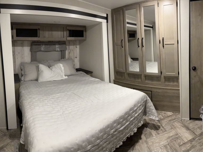 Comfortable RV rental bedroom with a queen bed, overhead cabinets, and a mirrored wardrobe. Ideal for travel accommodation.. Forest River Palomino Puma 2023