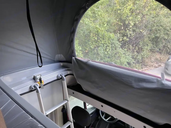 Interior of an RV rental pop-top camper, showing a ladder leading to a sleeping bunk and an outdoor view through the canvas window.. Winnebago Solis 59PX 2023