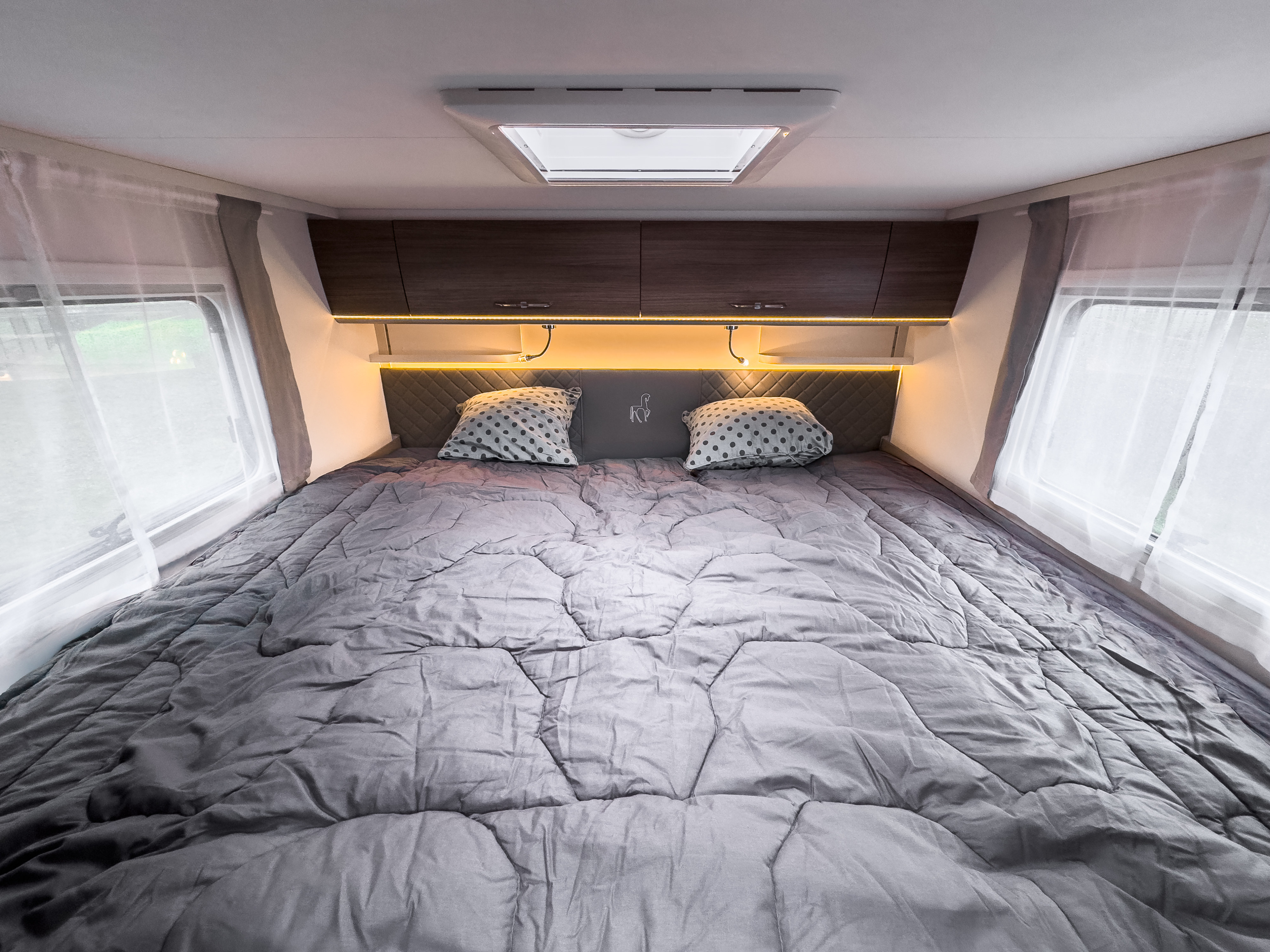 Experience comfort in this RV rental bedroom: a cozy bed with a grey comforter, ambient lighting, overhead storage, and large windows.. COSY COMFORT MC LOUIS 2024