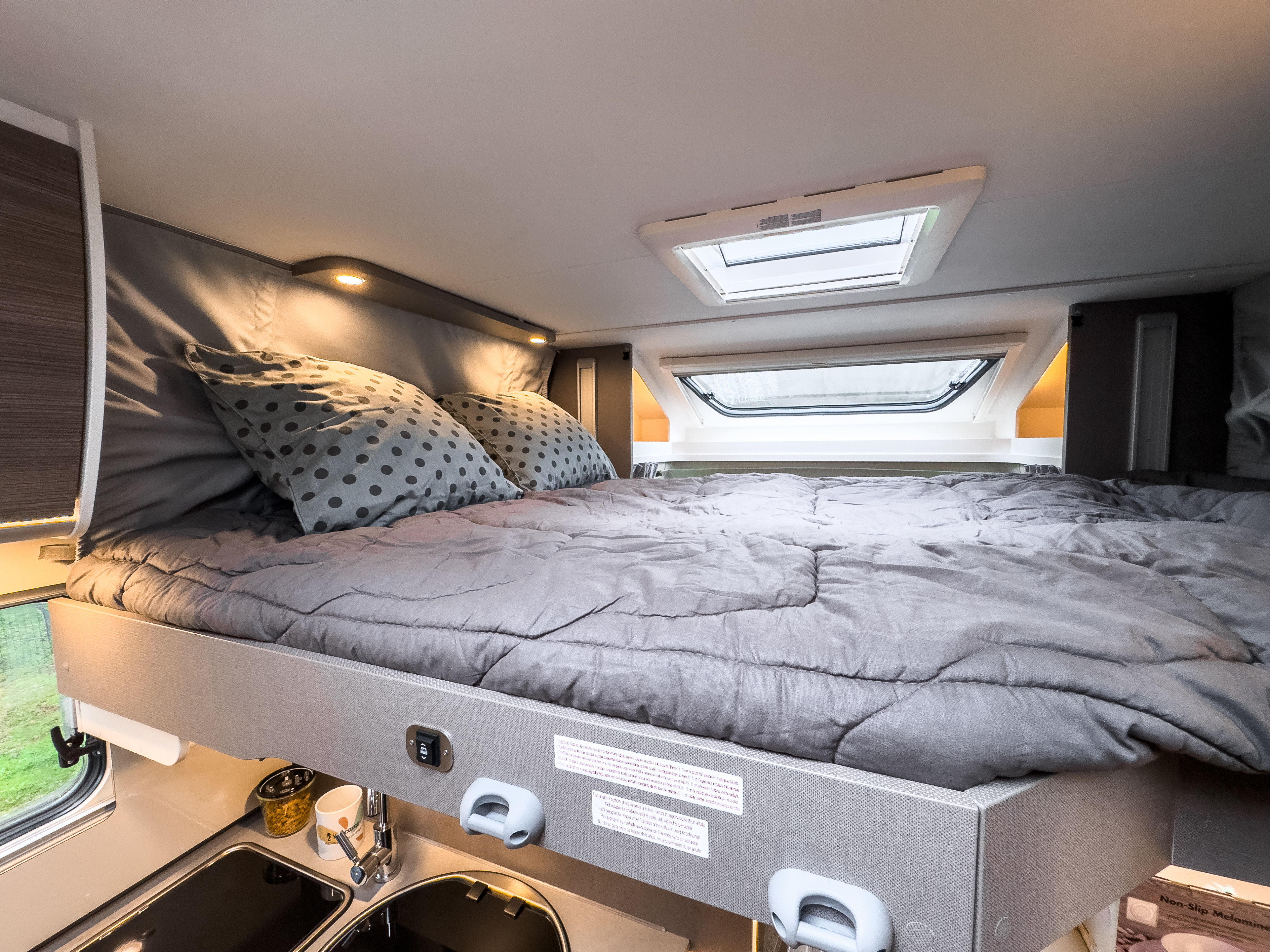 RV rental's cozy bed area with grey duvet and pillows, overhead lights, a skylight, and a compact kitchen below.. COSY COMFORT MC LOUIS 2024