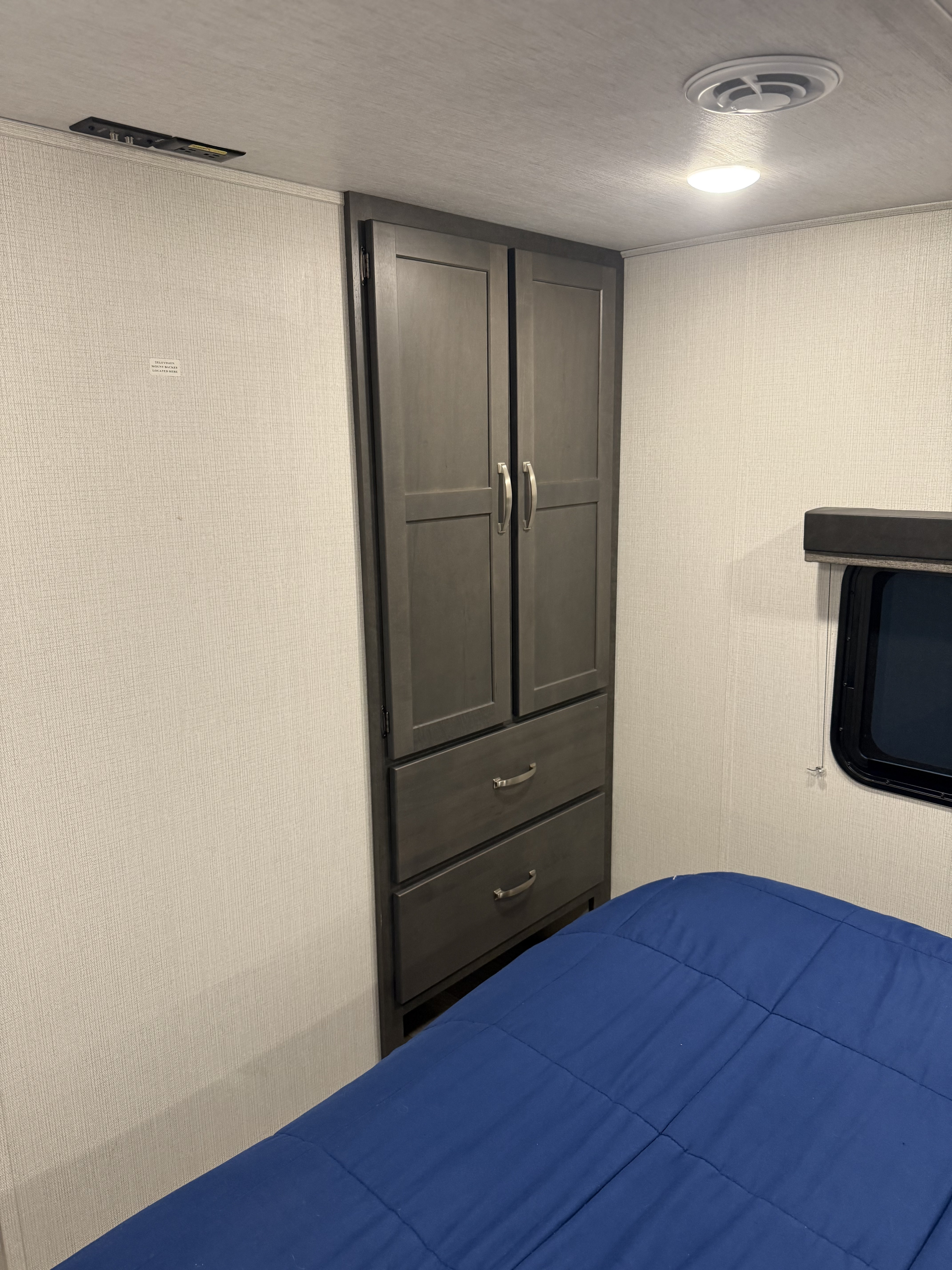 Inside an RV rental: A compact bedroom with a blue bed, built-in wardrobe, window, and TV mounting area.. Keystone RV Cougar 2024