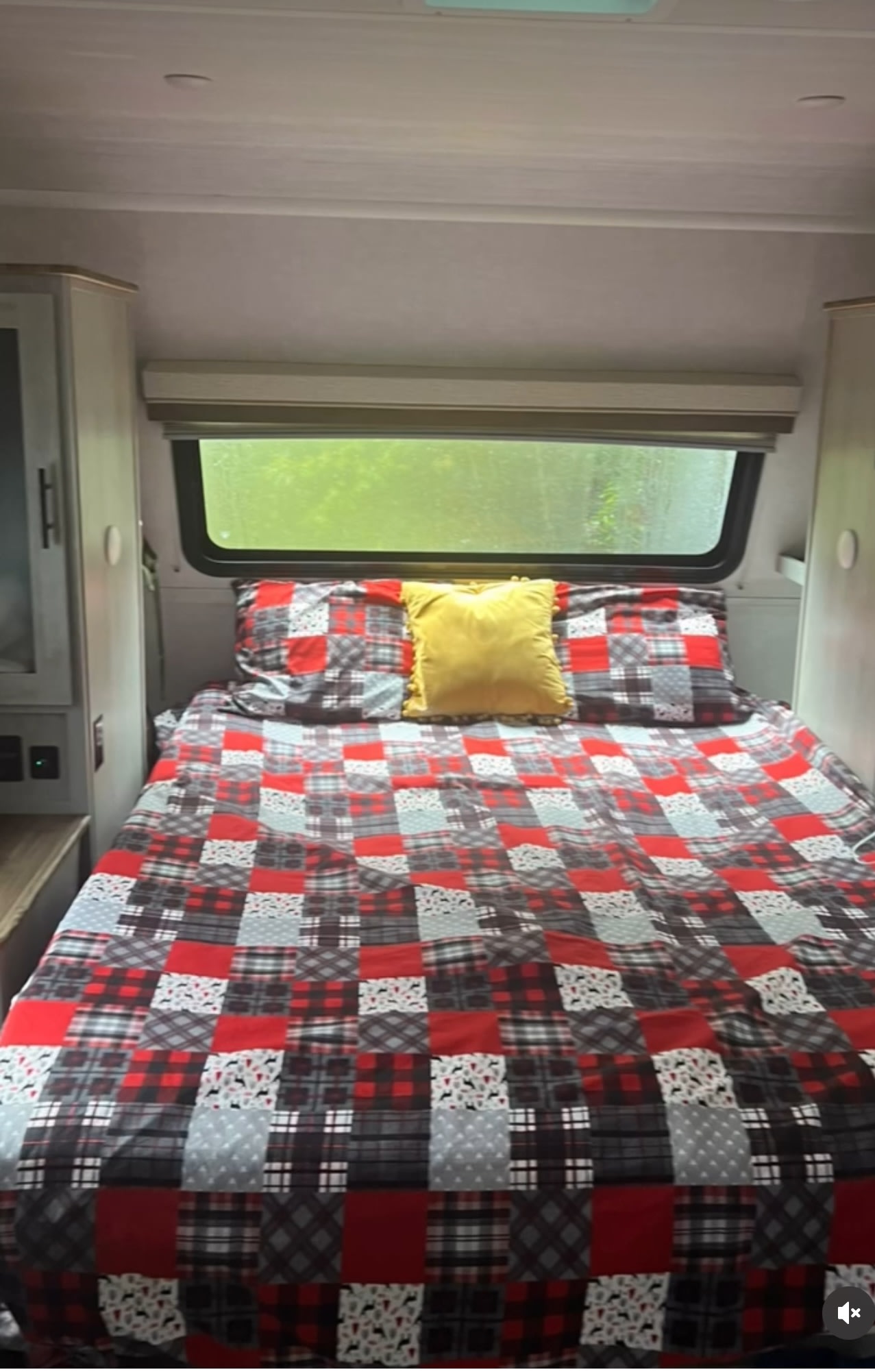 Inside a comfortable RV rental: a bed with a red plaid comforter, yellow accent pillow, and nature view from the window.. Forest River IBEXMBS 2023