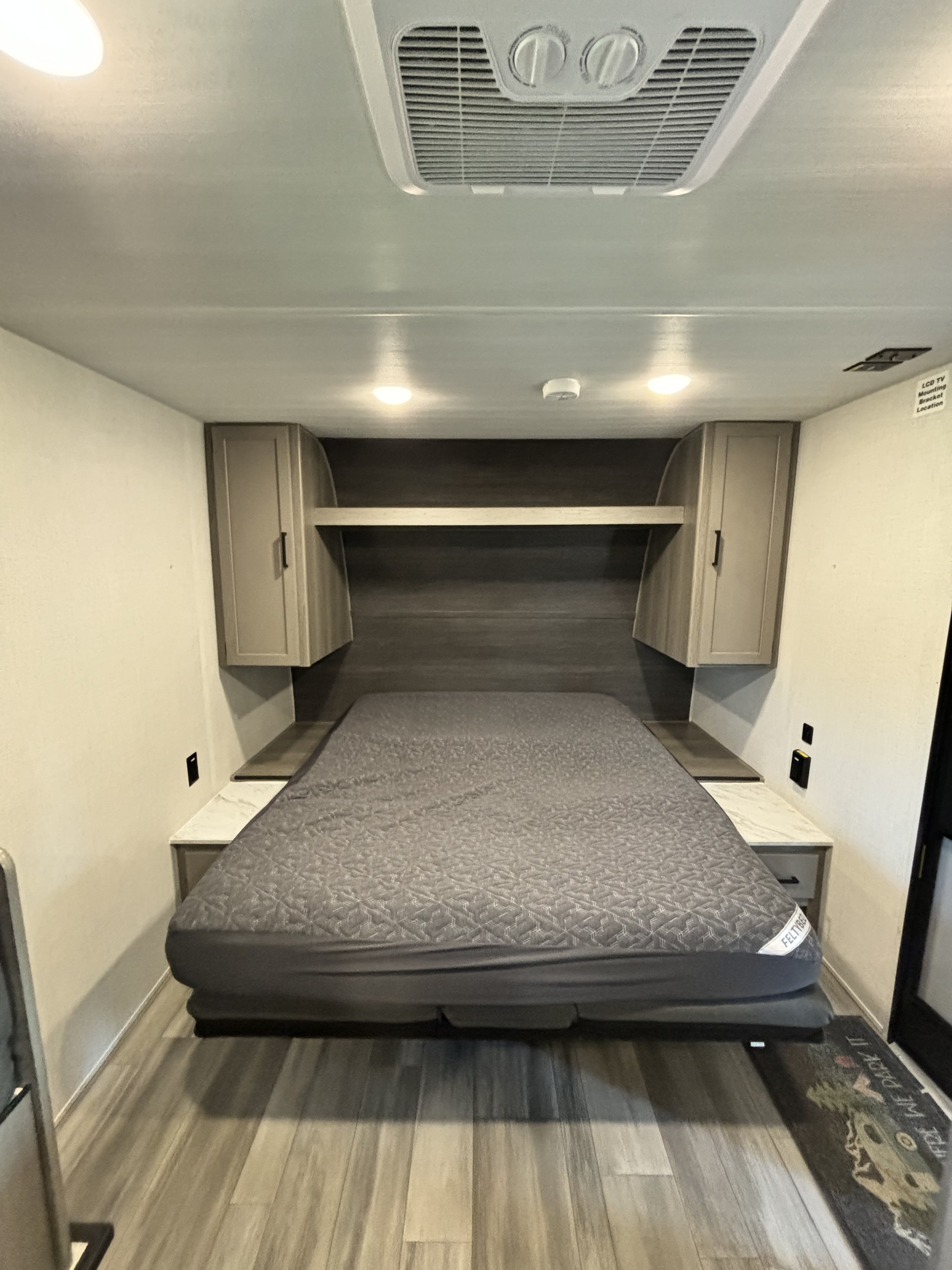 RV rental bedroom: a comfy grey bed, overhead cabinets, and built-in nightstands. Ceiling AC provides comfort in this compact space.. Keystone RV Hideout 181BH 2024