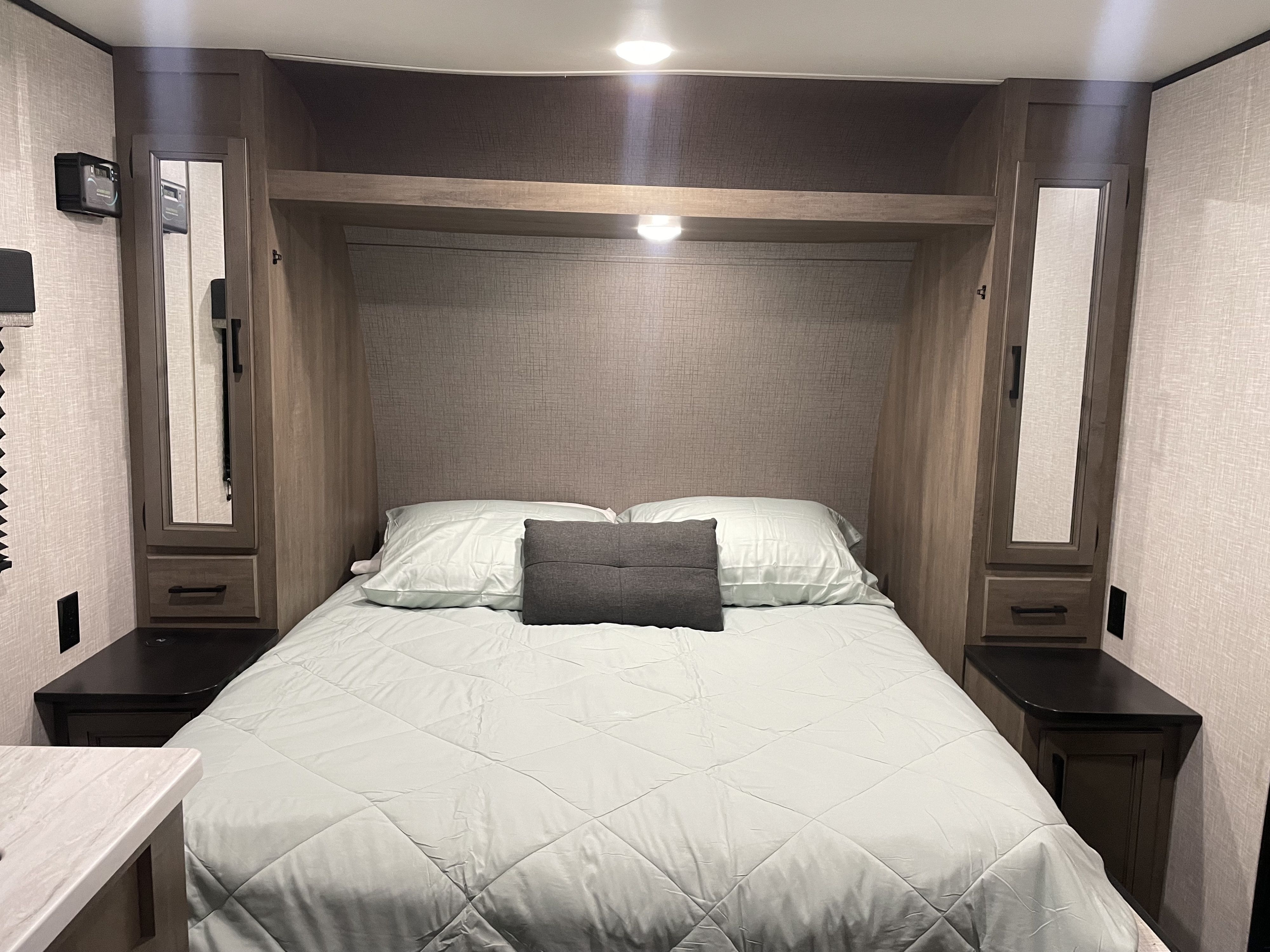 An RV rental bedroom features a cozy bed with light green bedding, flanked by mirrored storage cabinets, nightstands, and overhead lighting.. Jayco Autumn Ridge 20MB 2023