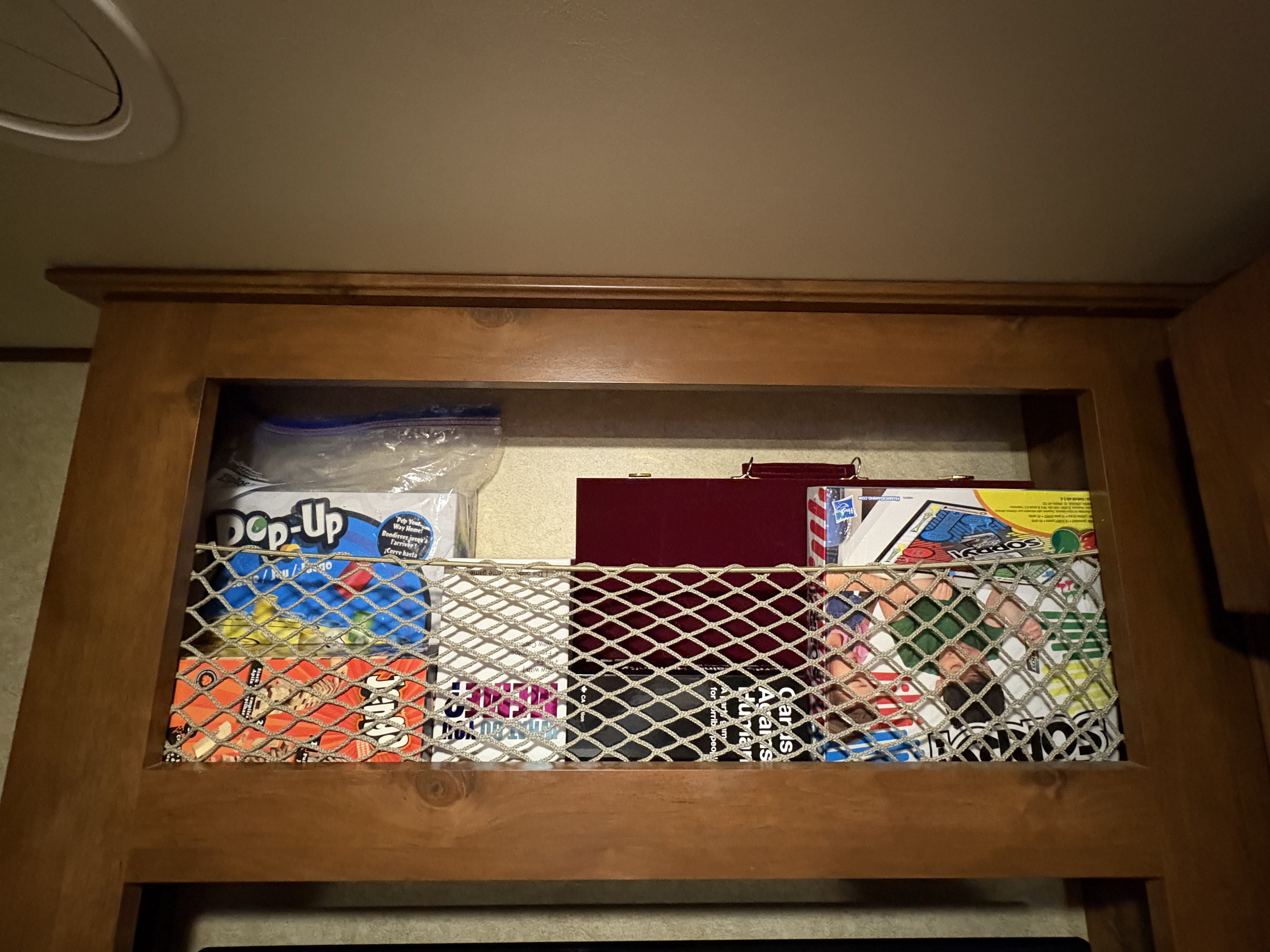 Overhead RV rental cabinet storing board games (Pop-Up, Sorry!) securely behind a mesh net for travel entertainment.. Open Range RV Light 2014