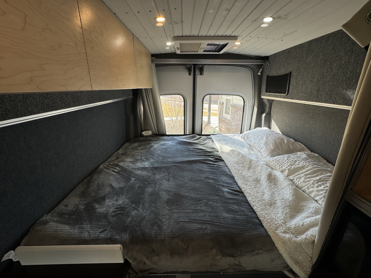 Cozy RV rental interior with a bed, overhead wood cabinets, and rear windows. Experience comfort in this camper van.. Ford Transit 2015