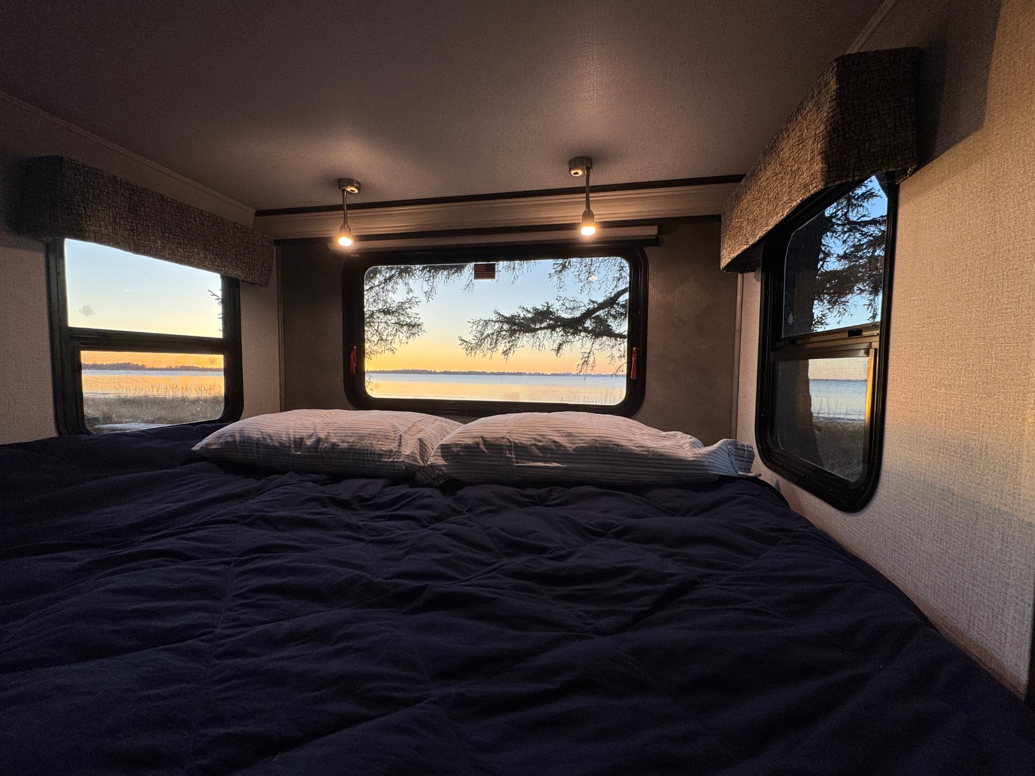 Inside an RV rental: a cozy bedroom with dark blue bedding, striped pillows, and a serene waterfront sunset view.. Forester TS 2020